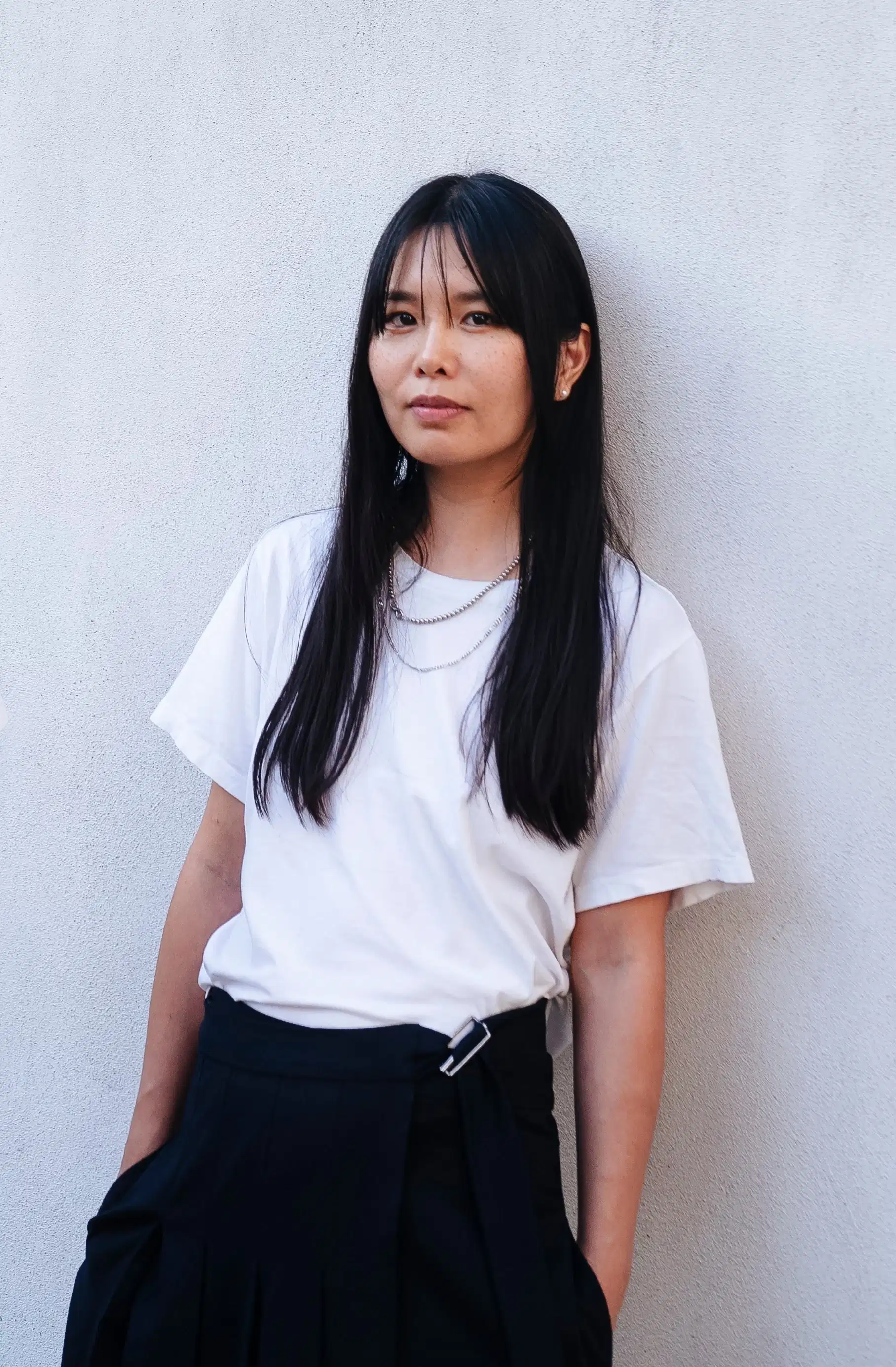 A photo of Ami Lien. She is leaning against a white wall and looking into the camera with a relaxed expression. She is wearing a white shirt and black pants and has two silver chain necklaces on.