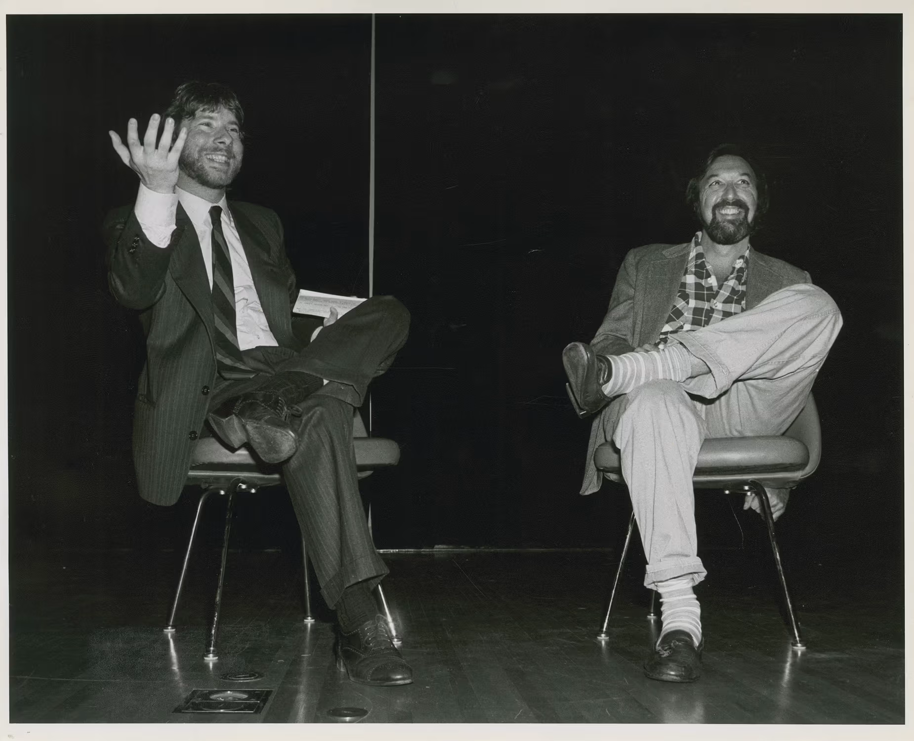 A black-and-white photo of two people sitting on chairs and smiling at something off-camera.