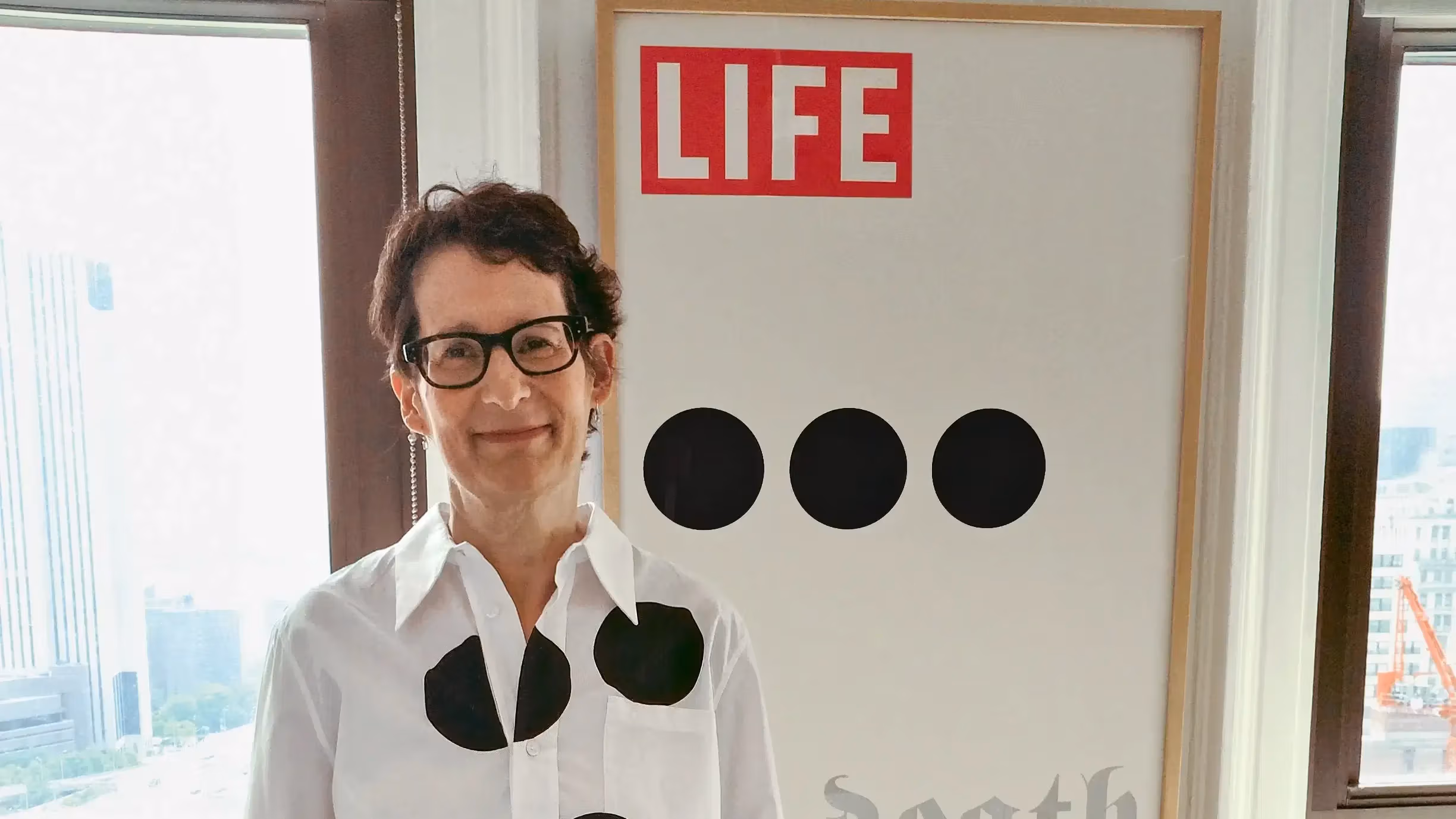 A photograph of a woman wearing a shirt with large polka dots and standing in front of a poster showing similarly-sized dots. 