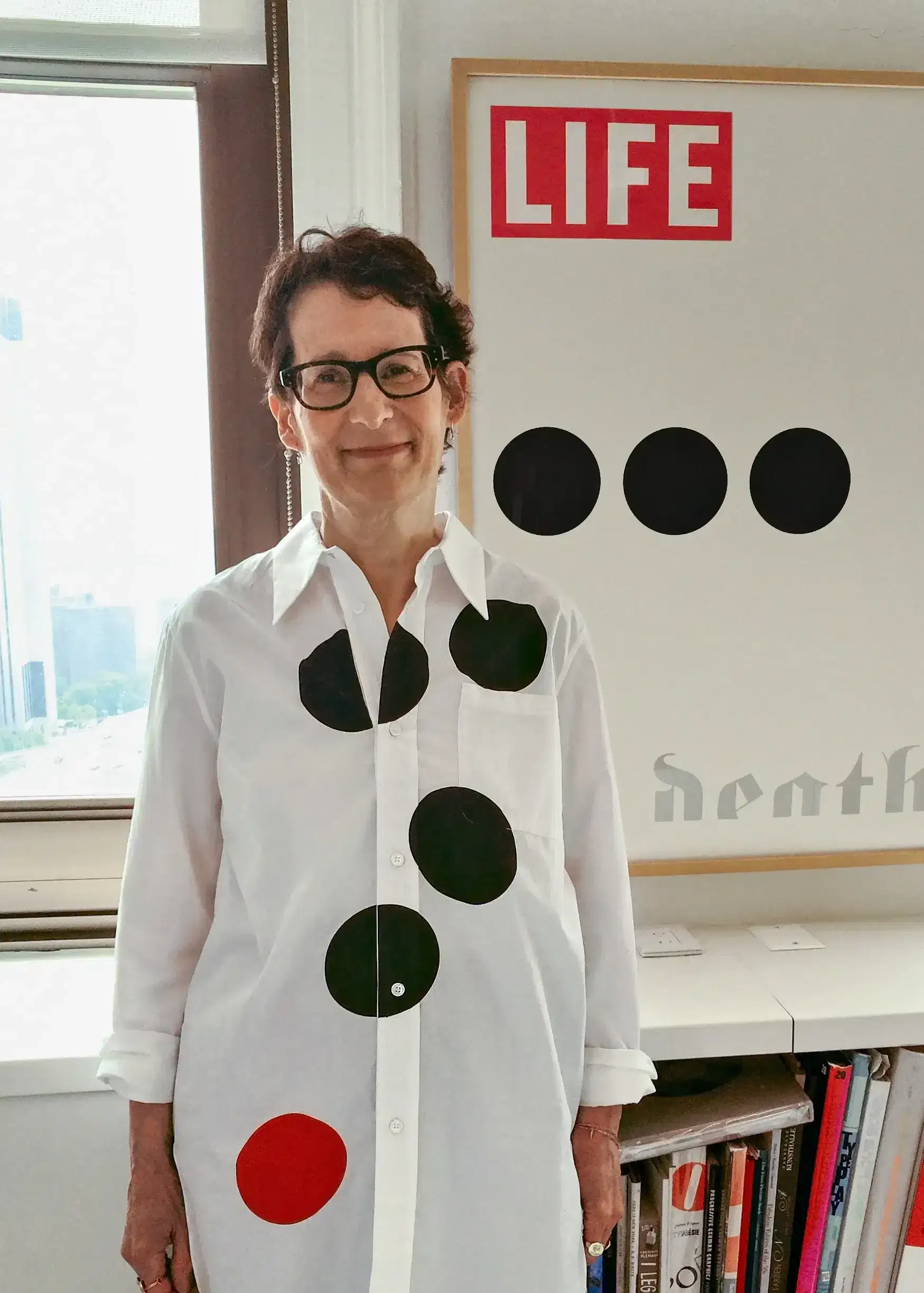 A photograph of a woman wearing a shirt with large polka dots and standing in front of a poster showing similarly-sized dots. 