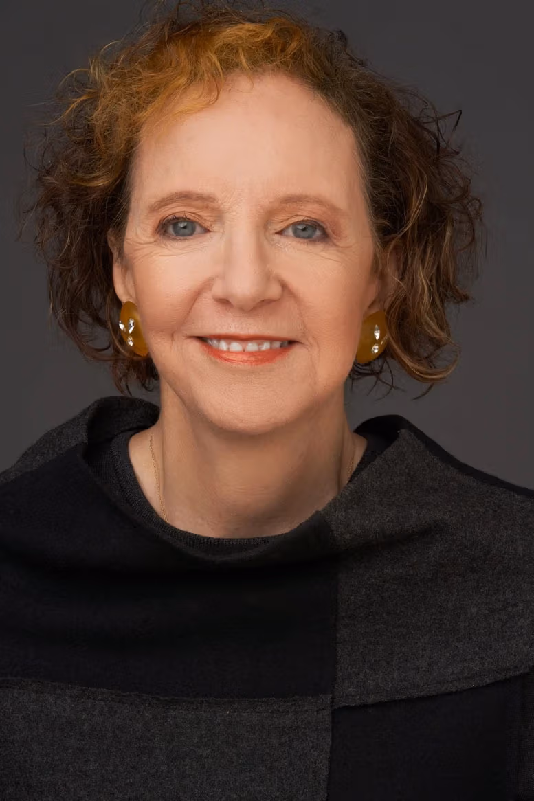 A photograph of Suzaan Boettger by Timothy Greenfield-Sanders. Suzaan is wearing a grey and black checkered sweater. She is smiling into the camera. The background is a solid grey color.