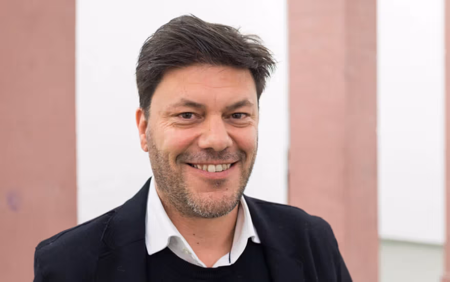 a smiling person with short brown hair and a black blazer on stands in front of a pink and white building exterior 