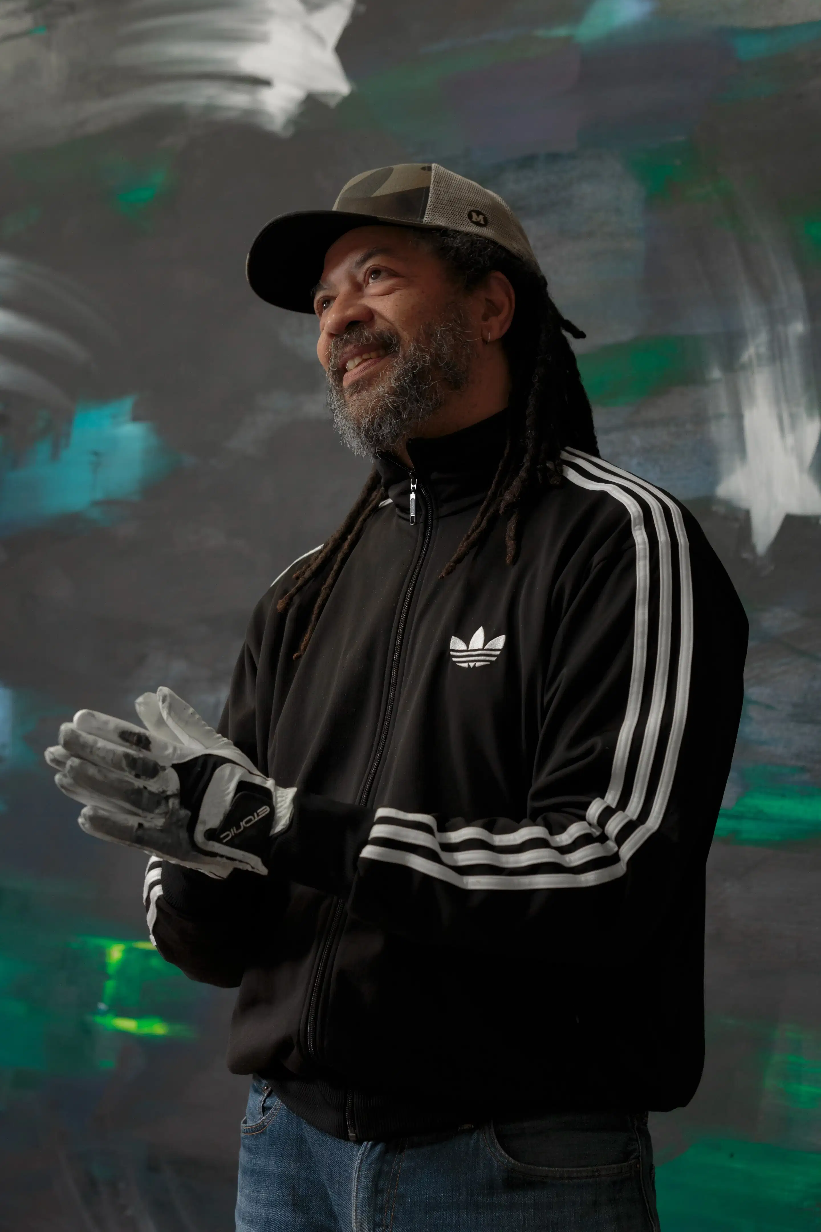 A man in a black jacket and hat stands smiling as he presses his hands together.