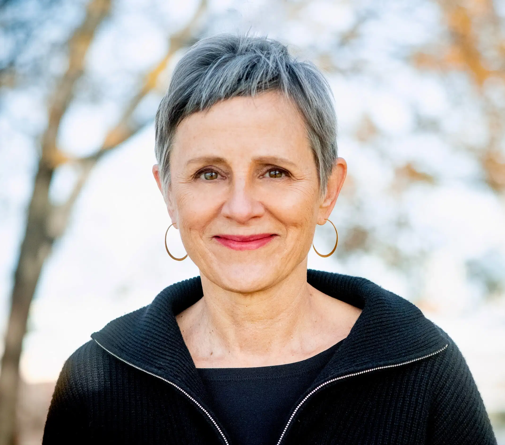 Susan Hapgood headshot. Susan is wearing a black sweater and has short grey and black hair. She is wearing thin hoop earrings and is smiling at the camera. In the background is a blurred natural scenery.
