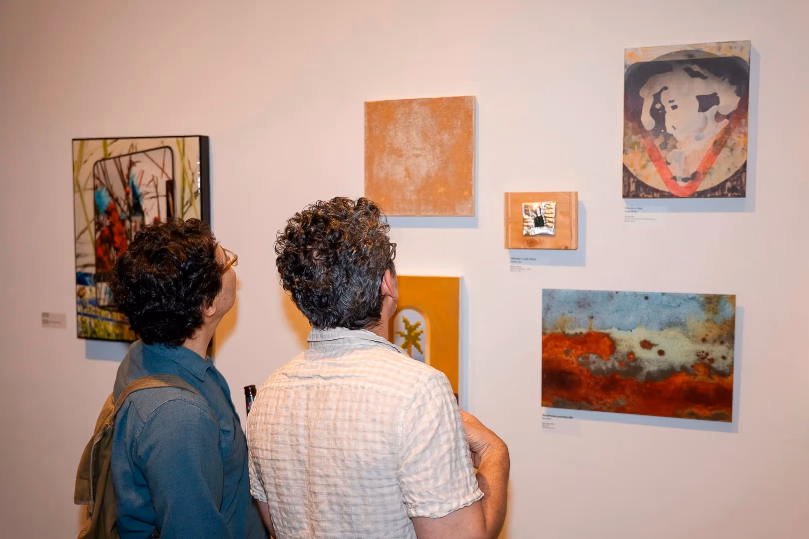 a person in a blue shirt and a person in a white shirt look at a wall of paintings
