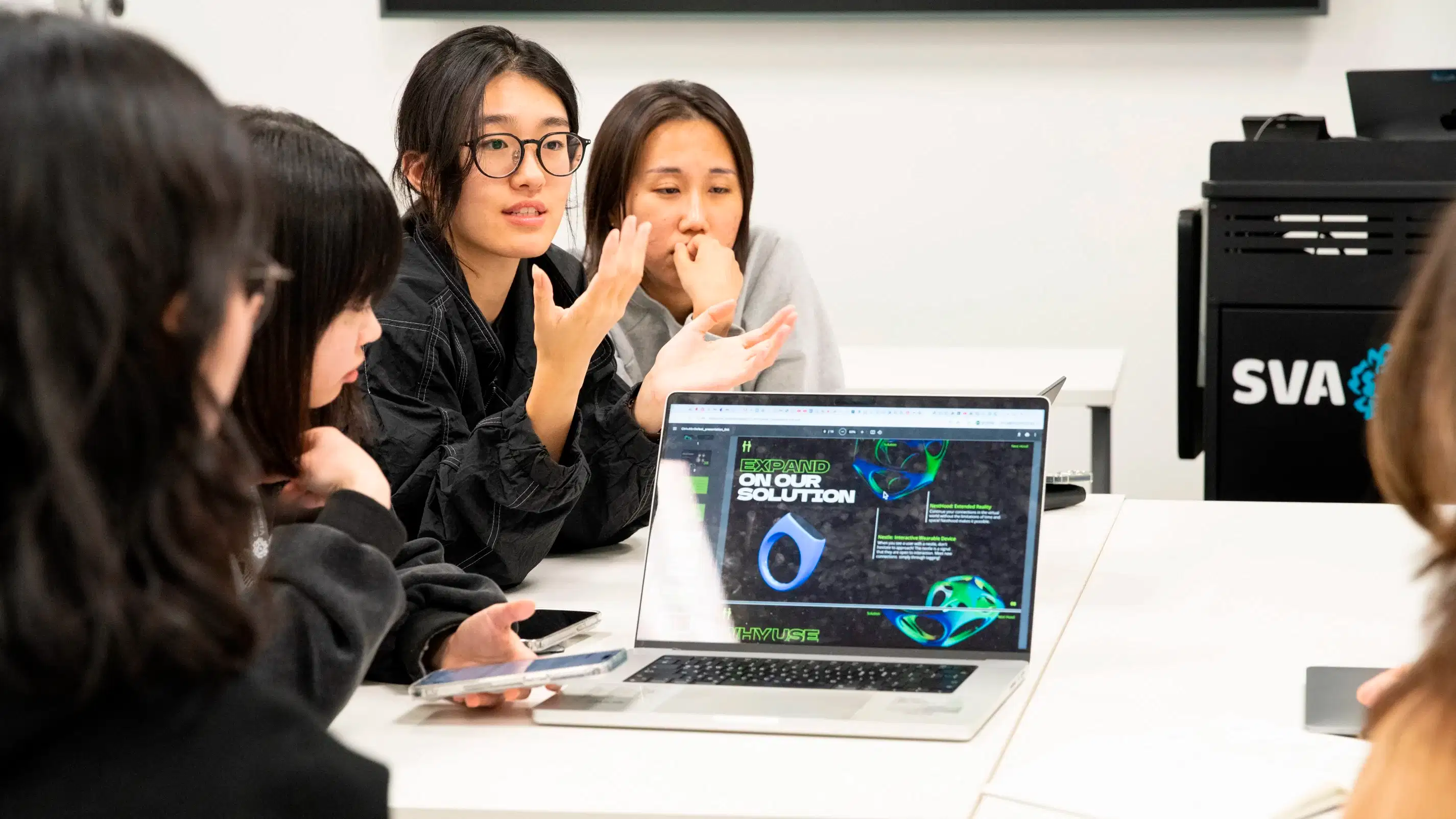 Four students sit at a table and present their work behind a laptop screen showing their presentation.