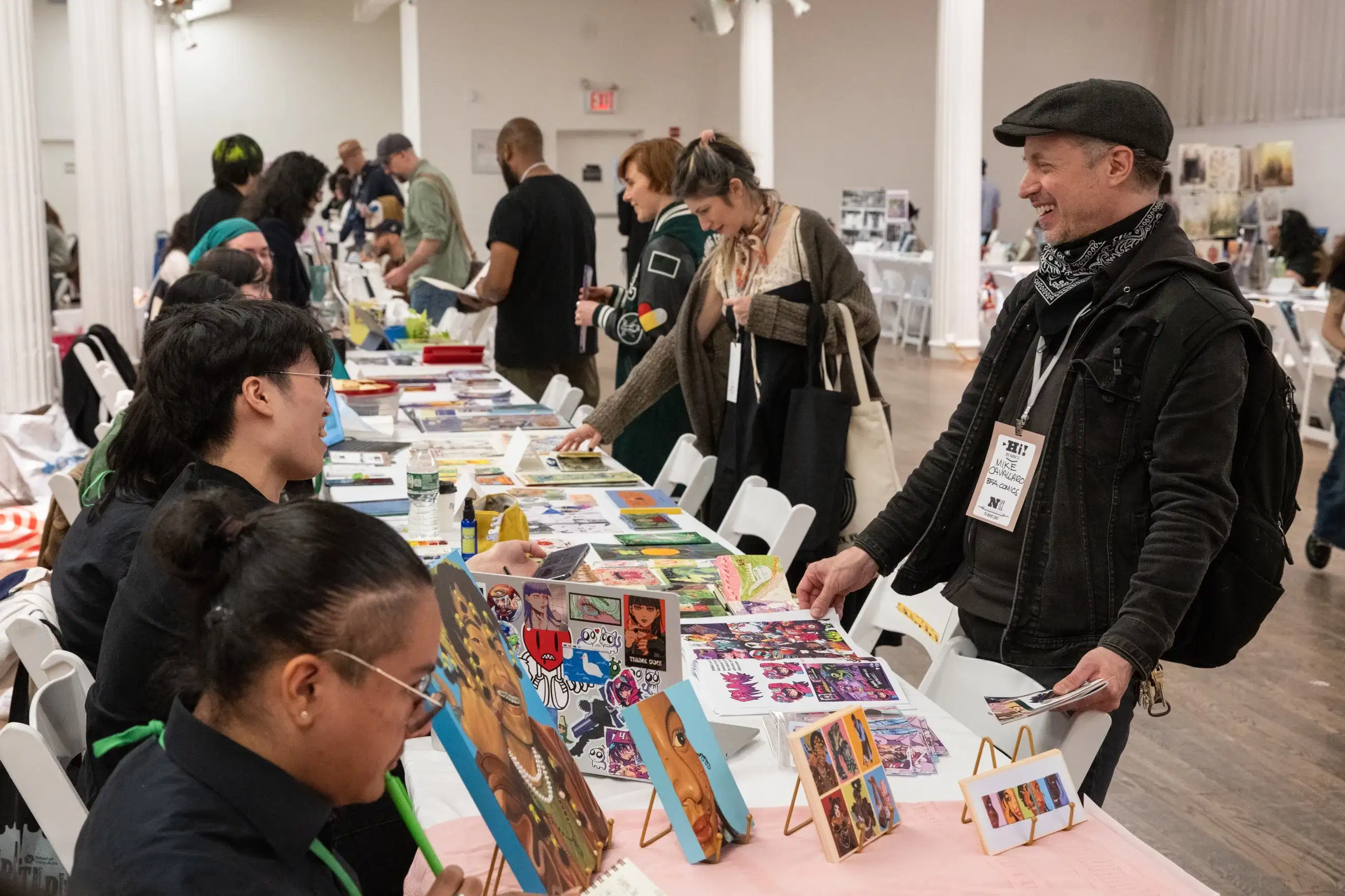 Students seated at a long table behind examples of their work, chatting with guests.