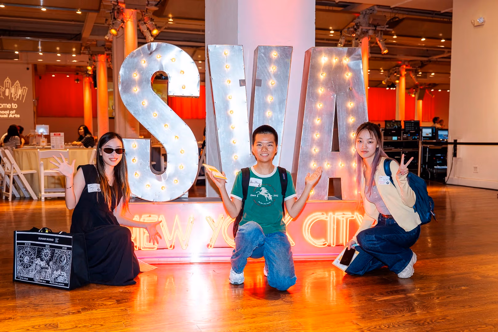 Three students pose in front of a large illuminated “SVA New York City” sign at an indoor welcome event.