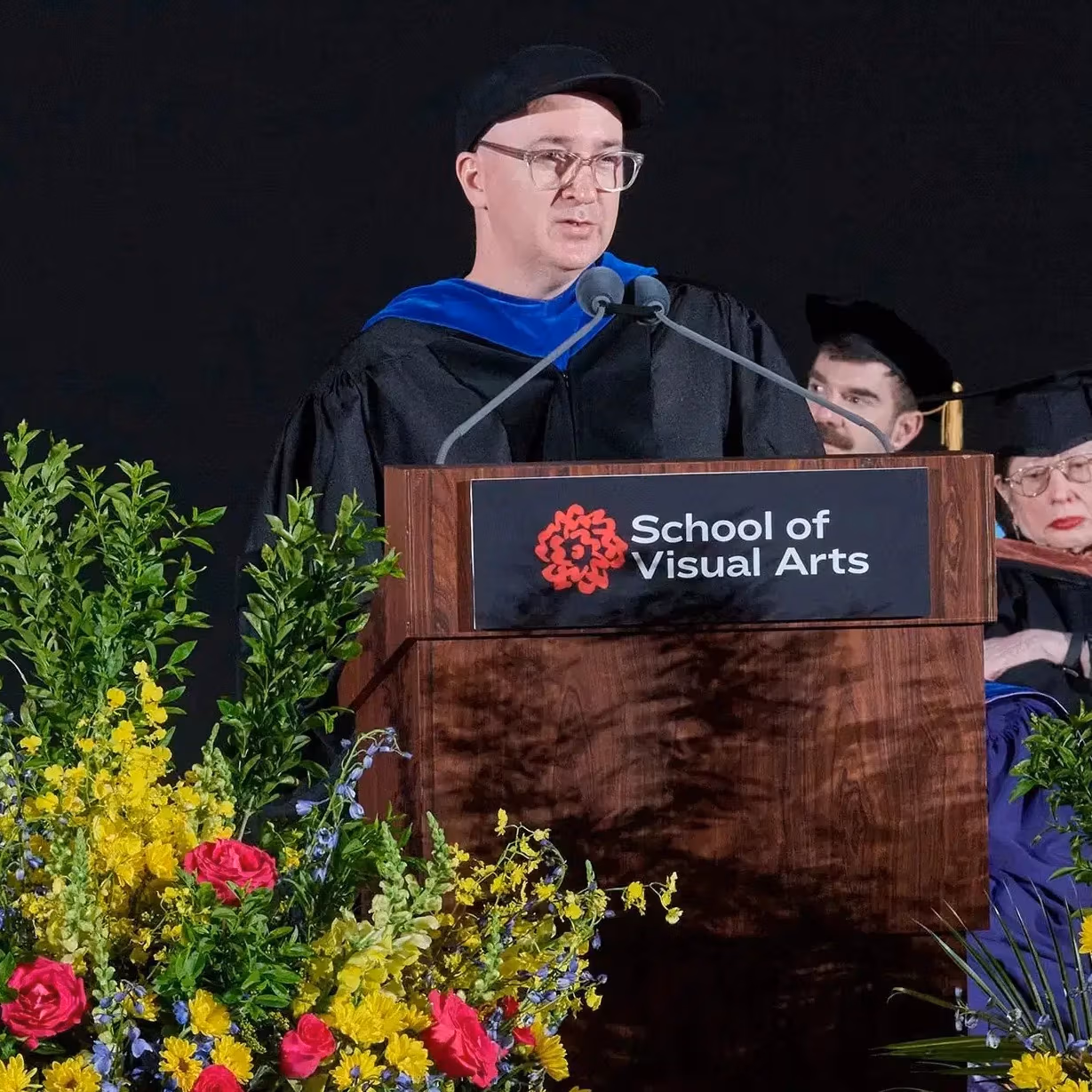 Speaker in academic regalia and a black cap stands at a podium with School of Visual Arts signage, speaking into a microphone behind floral arrangements.