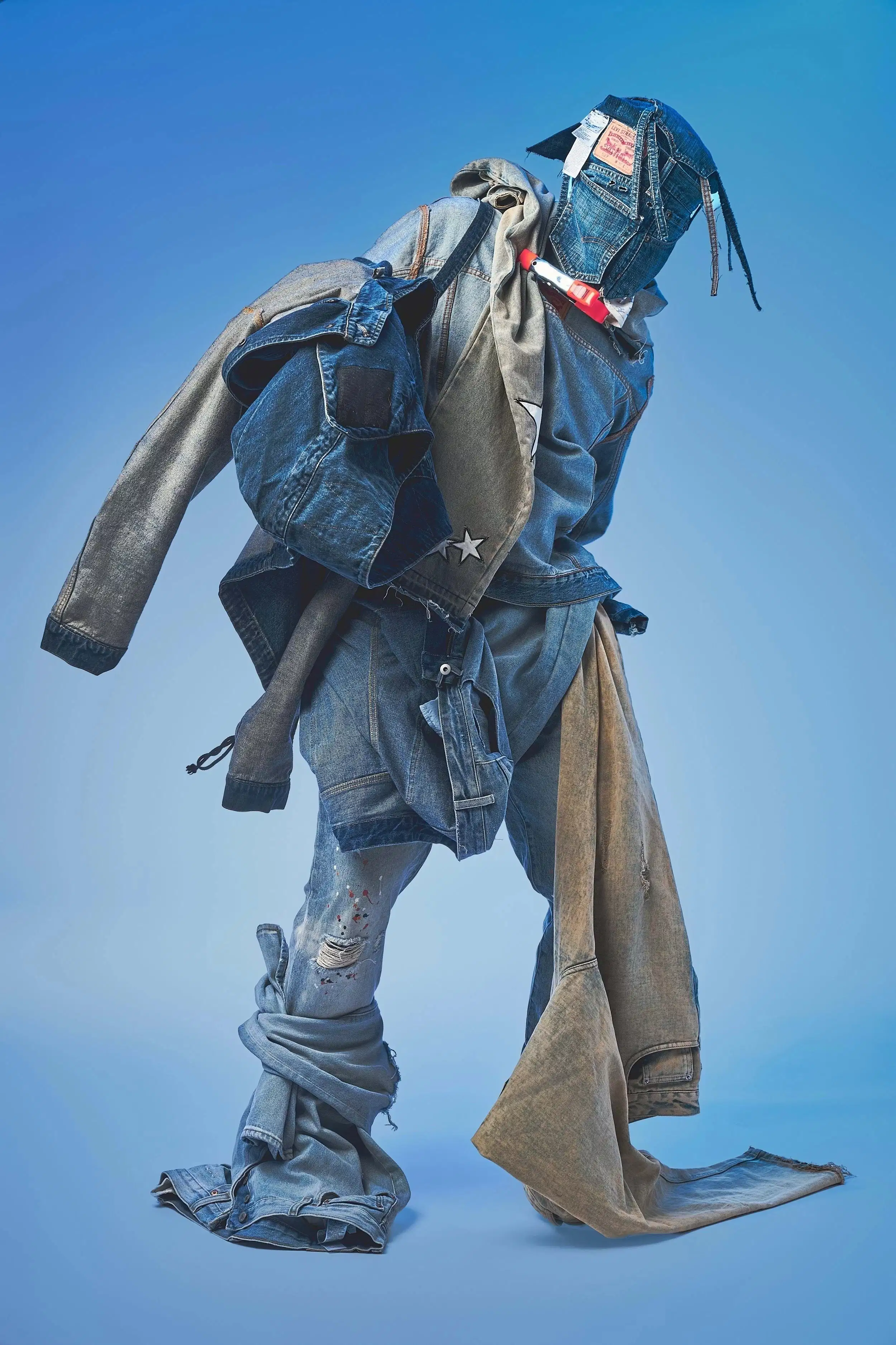 A studio fashion portrait of a model covered in an all-denim blue outfit and wearing a mask made of denim. The model stands in front of a blue background.