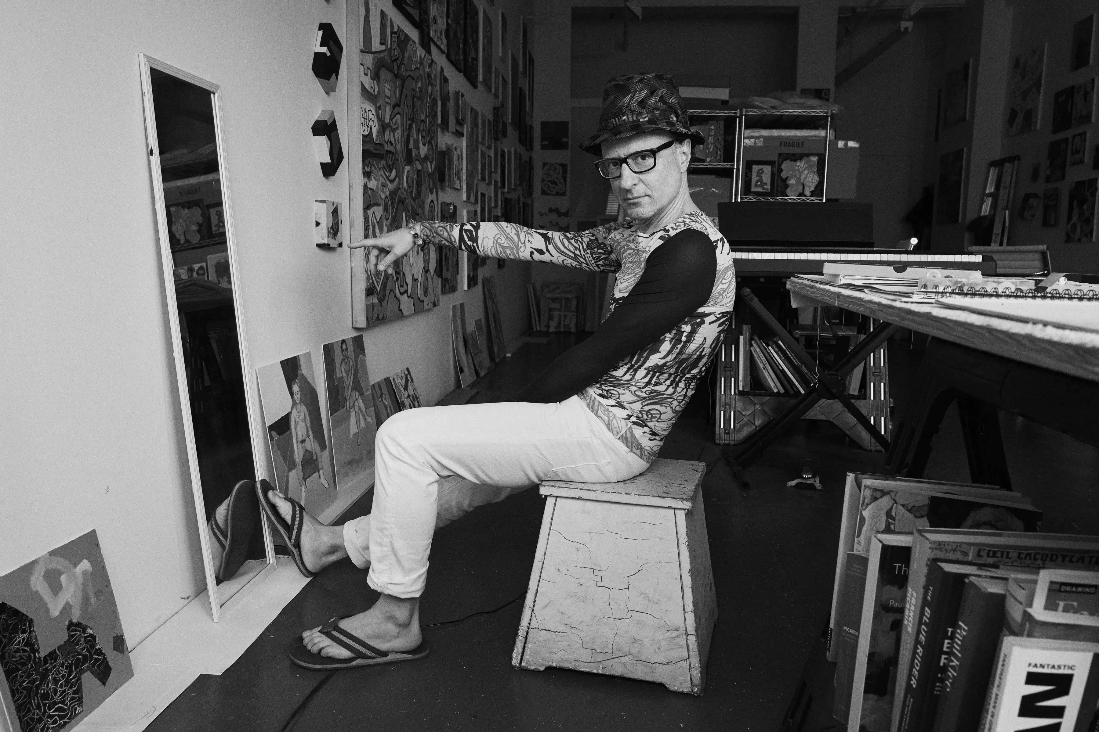 Black and white photo portrait of the artist Wayne Koestenbaum pointing at a painting in his studio.