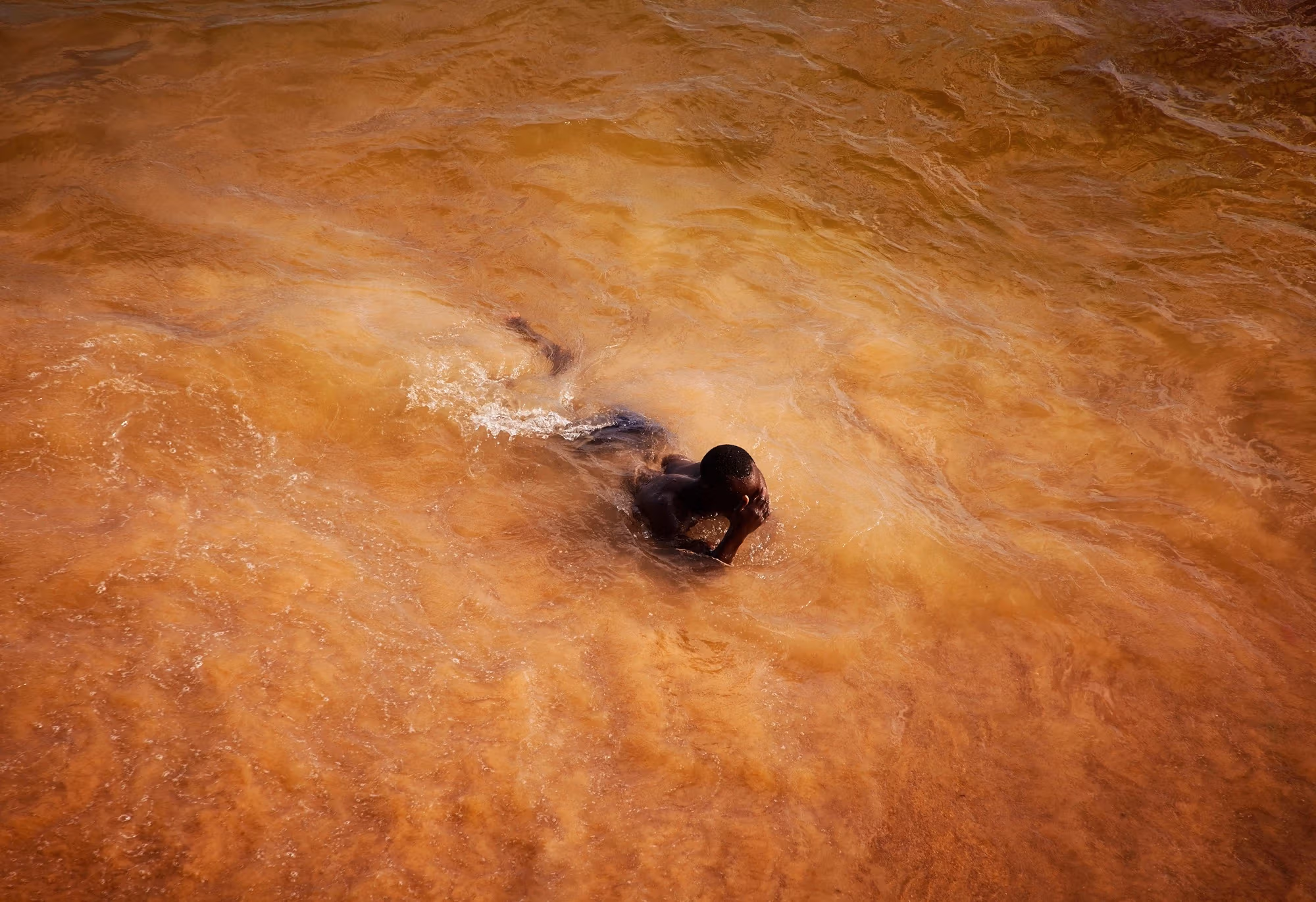 In this color photograph, a man emerges from the ocean, which has taken on a vibrant orange-ish cast in rural Saly, Senegal.
