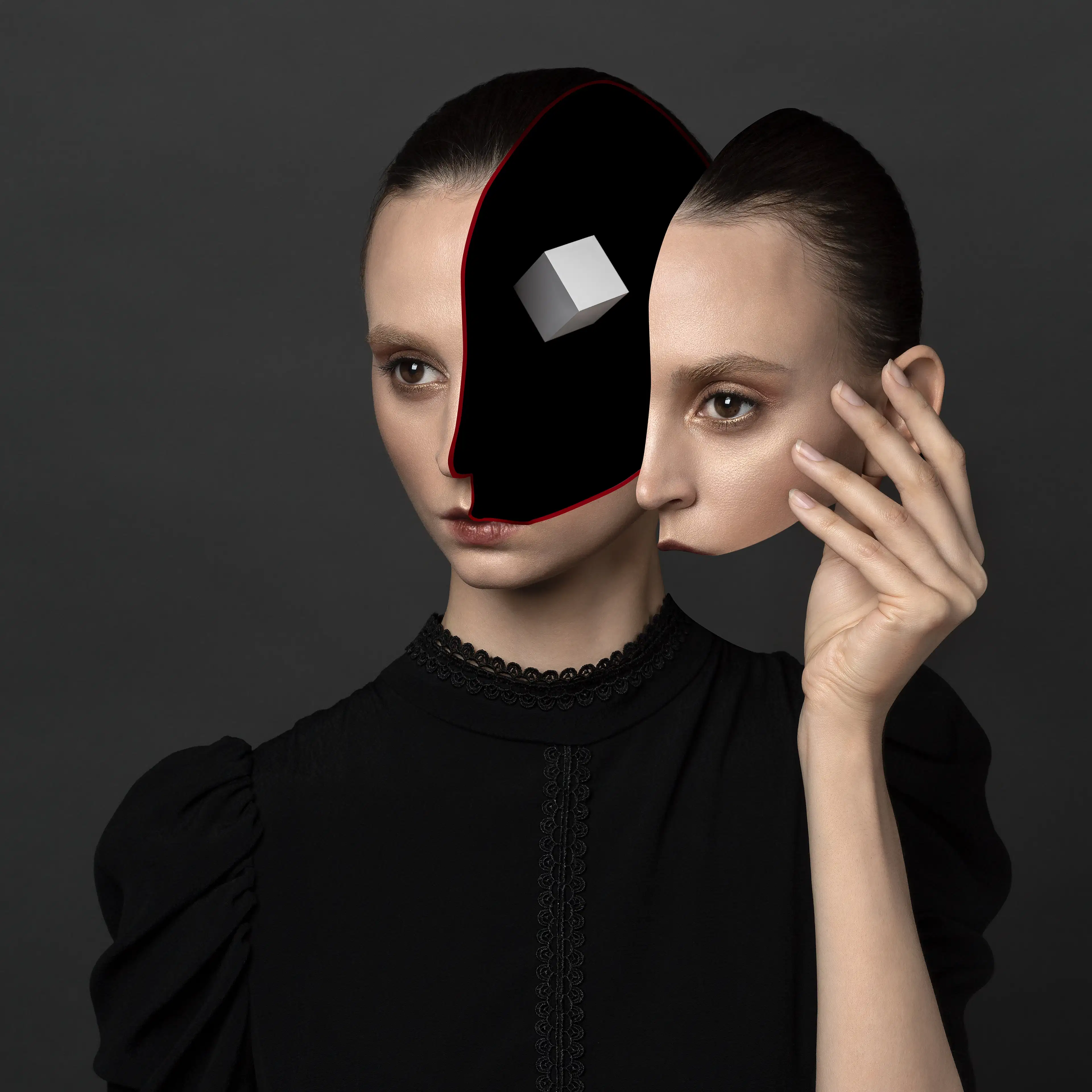 A studio headshot portrait of a woman against a gray background, wearing a black blouse. The left portion of her face from forehead to mouth has been removed, leaving a black negative void. Suspended in the void is a small, white cube. The woman’s right hand is held up next to her head, and holding the removed portion of her face like a mask.