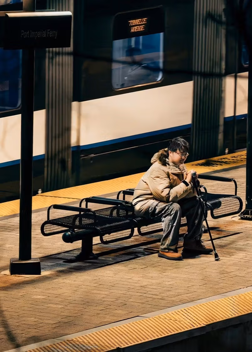 a person sits on a bench on an outdoor subway platform in a coat while holding a cane 