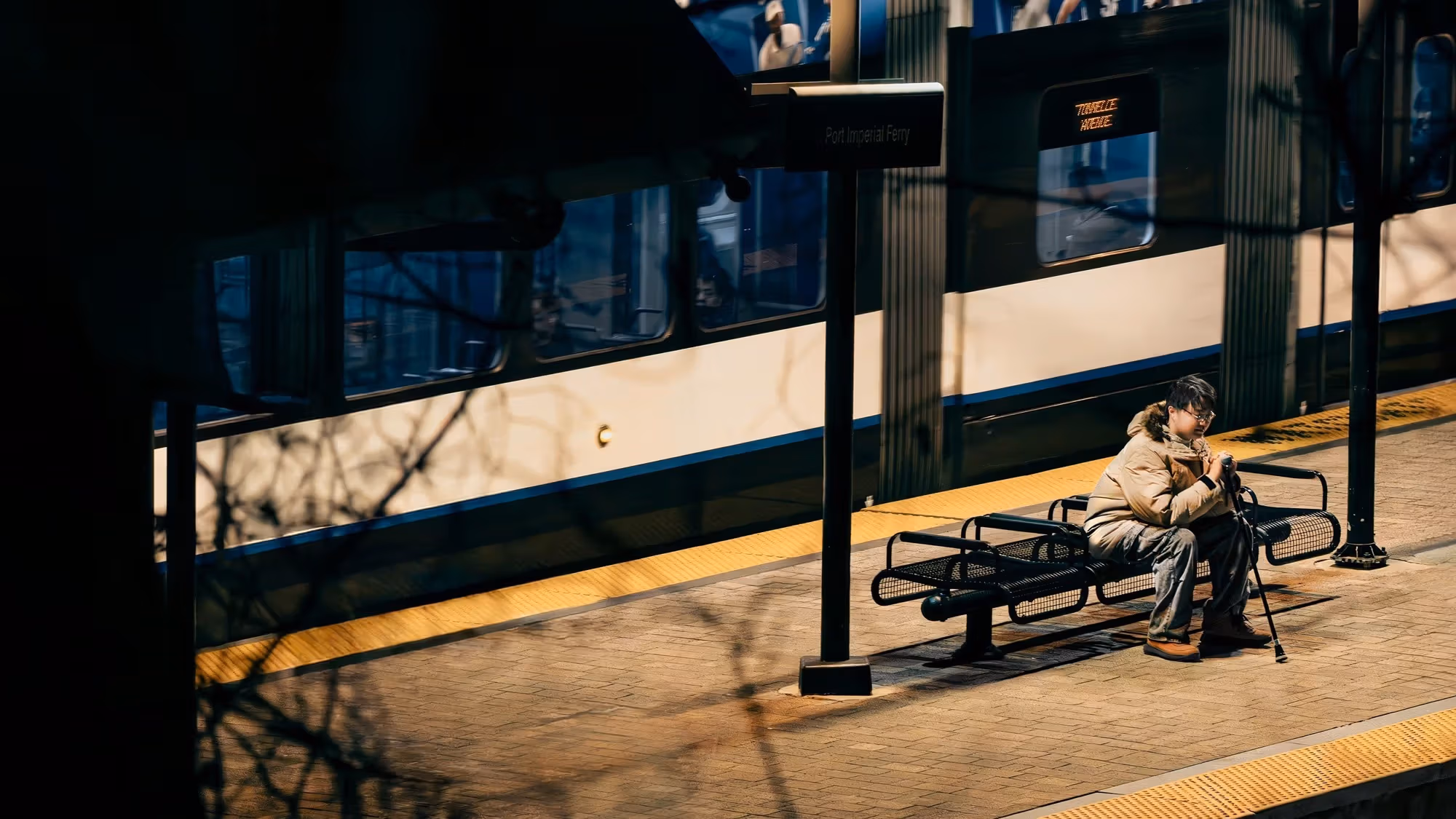 a person sits on a bench on an outdoor subway platform in a coat while holding a cane 