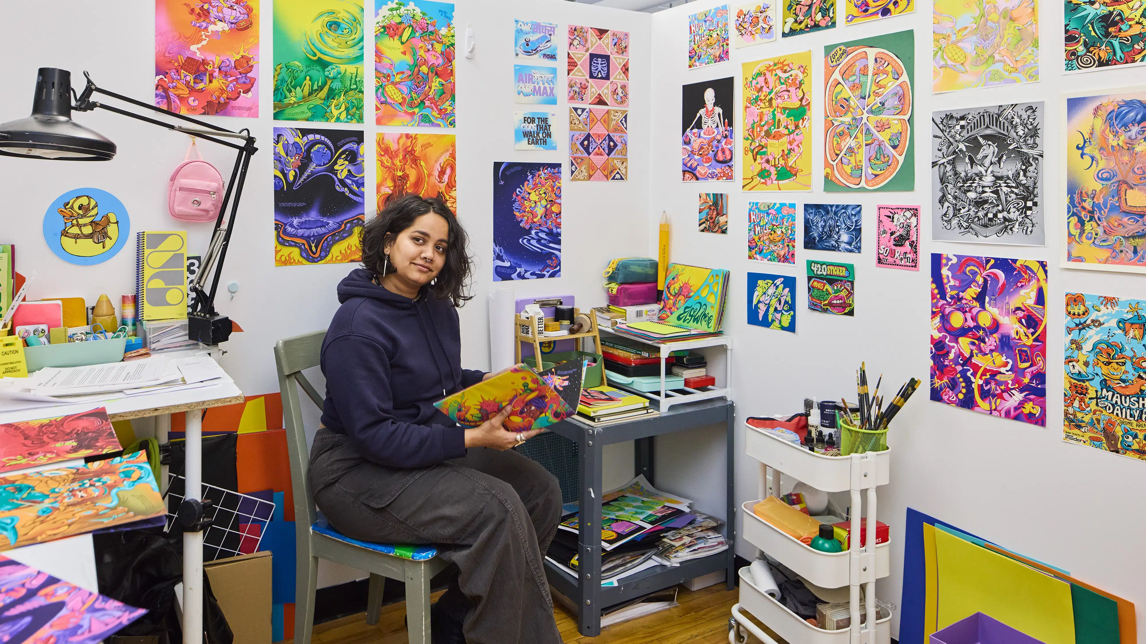 Female Illustration student holding a book in her personal studio space surrounded by her own vivid paintings covering the walls on display