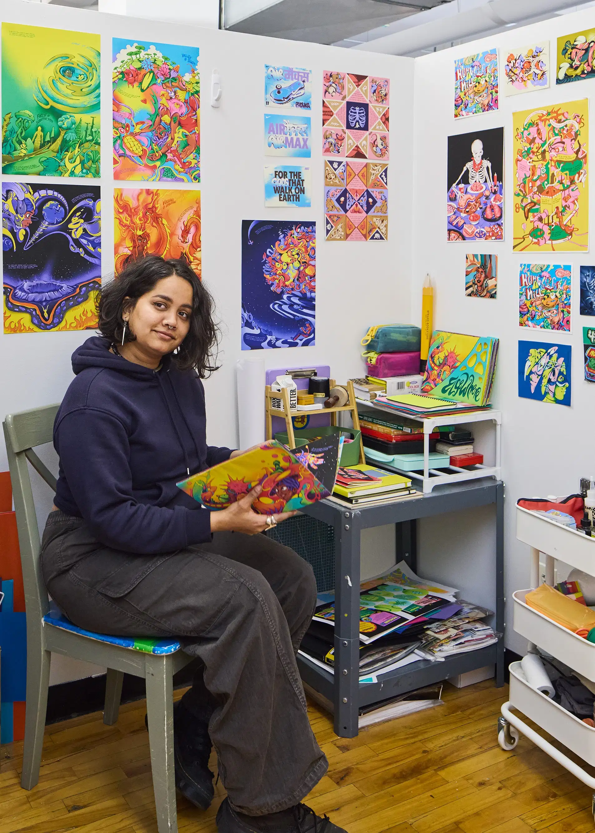 Female Illustration student holding a book in her personal studio space surrounded by her own vivid paintings covering the walls on display