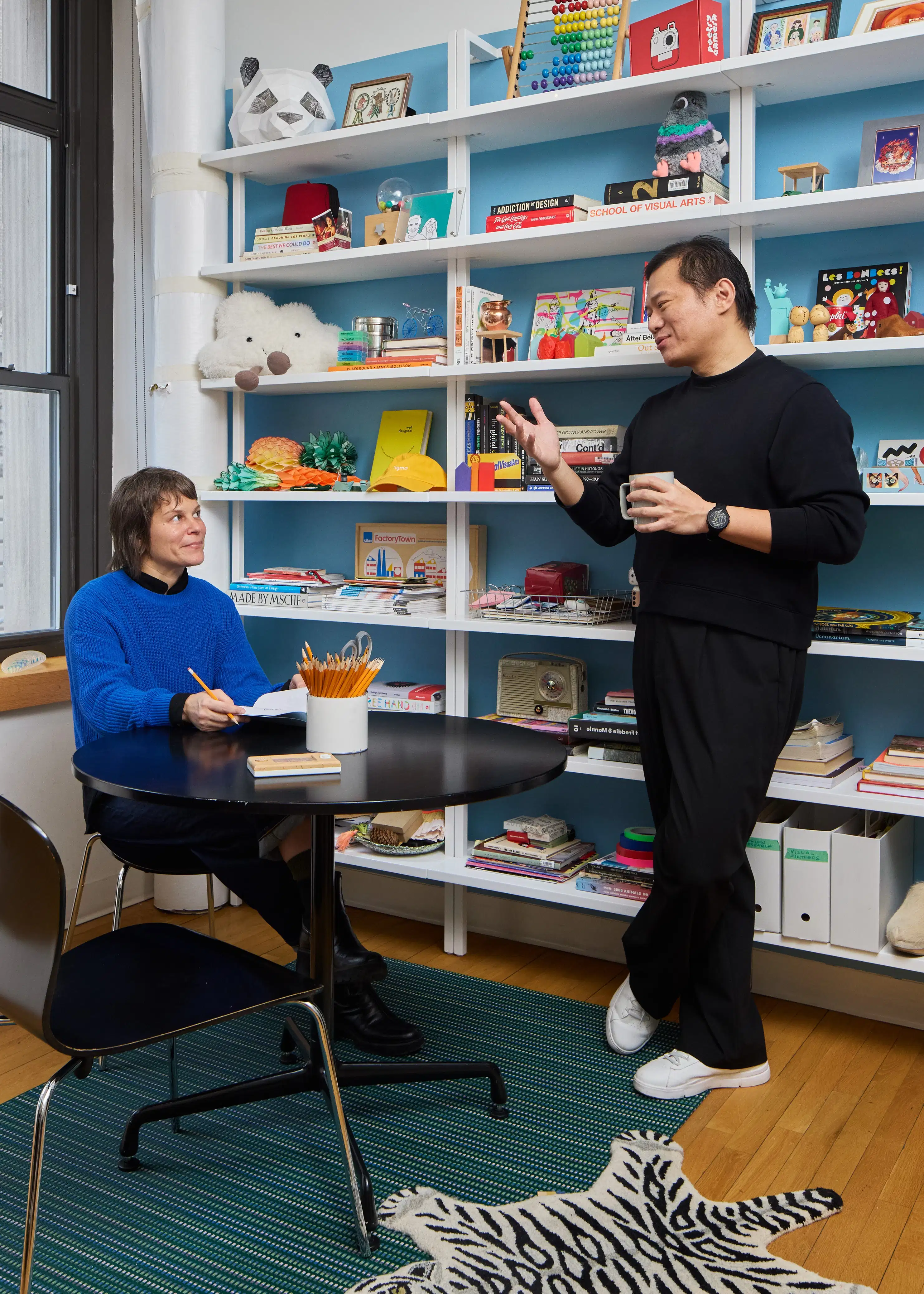 Male student standing in front of wall of shelves containing artifacts, art books and sculptural objects discussing ideas with female teacher sitting at a black table
