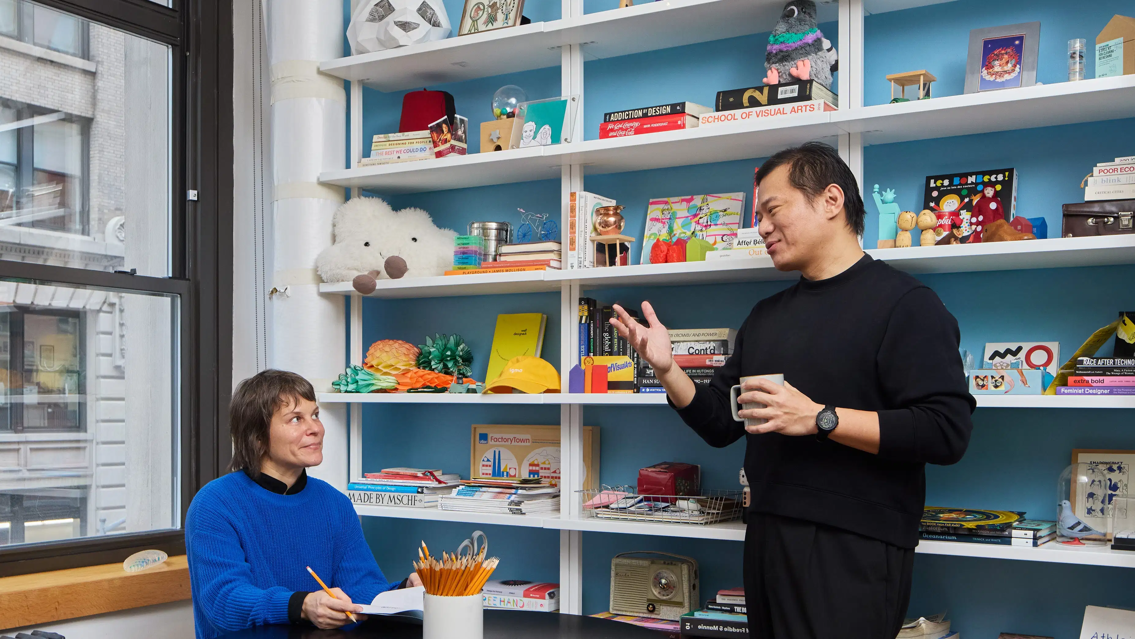 Male student standing in front of wall of shelves containing artifacts, art books and sculptural objects discussing ideas with female teacher sitting at a black table