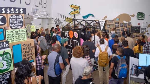 A crowd of people stands in a gallery space, gathered around a central art installation with colorful protest-style signs on the walls.