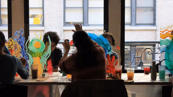 Artists paint on lucite panels while sitting down at a work table.