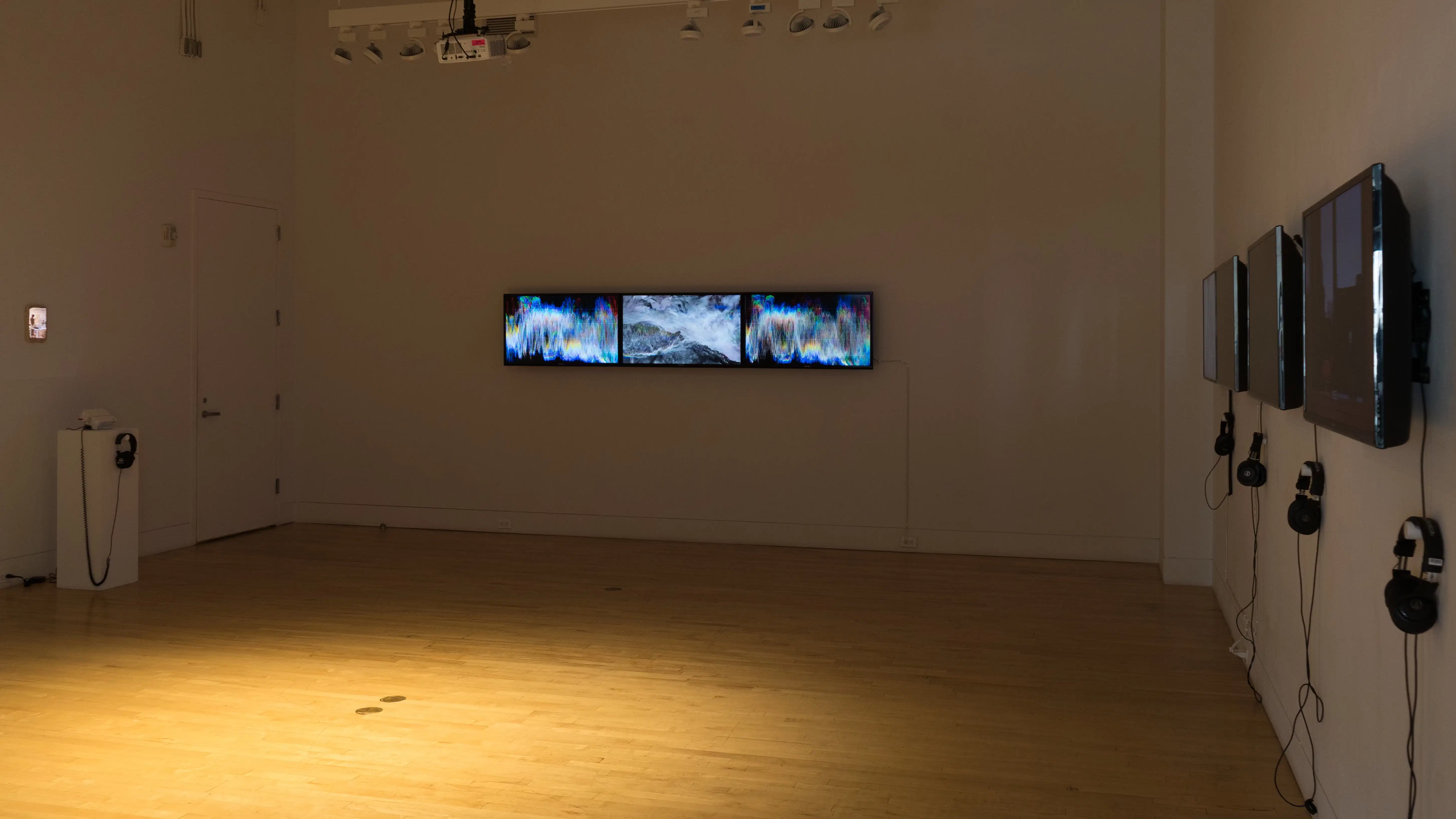 View of a gallery with three screens hung edge-to-edge in a horizontal line, with abstract video work playing, on the far back wall. On the left is a small projection on a mixed media surface, and on the far right are a line of monitors with their screens not fully visible.