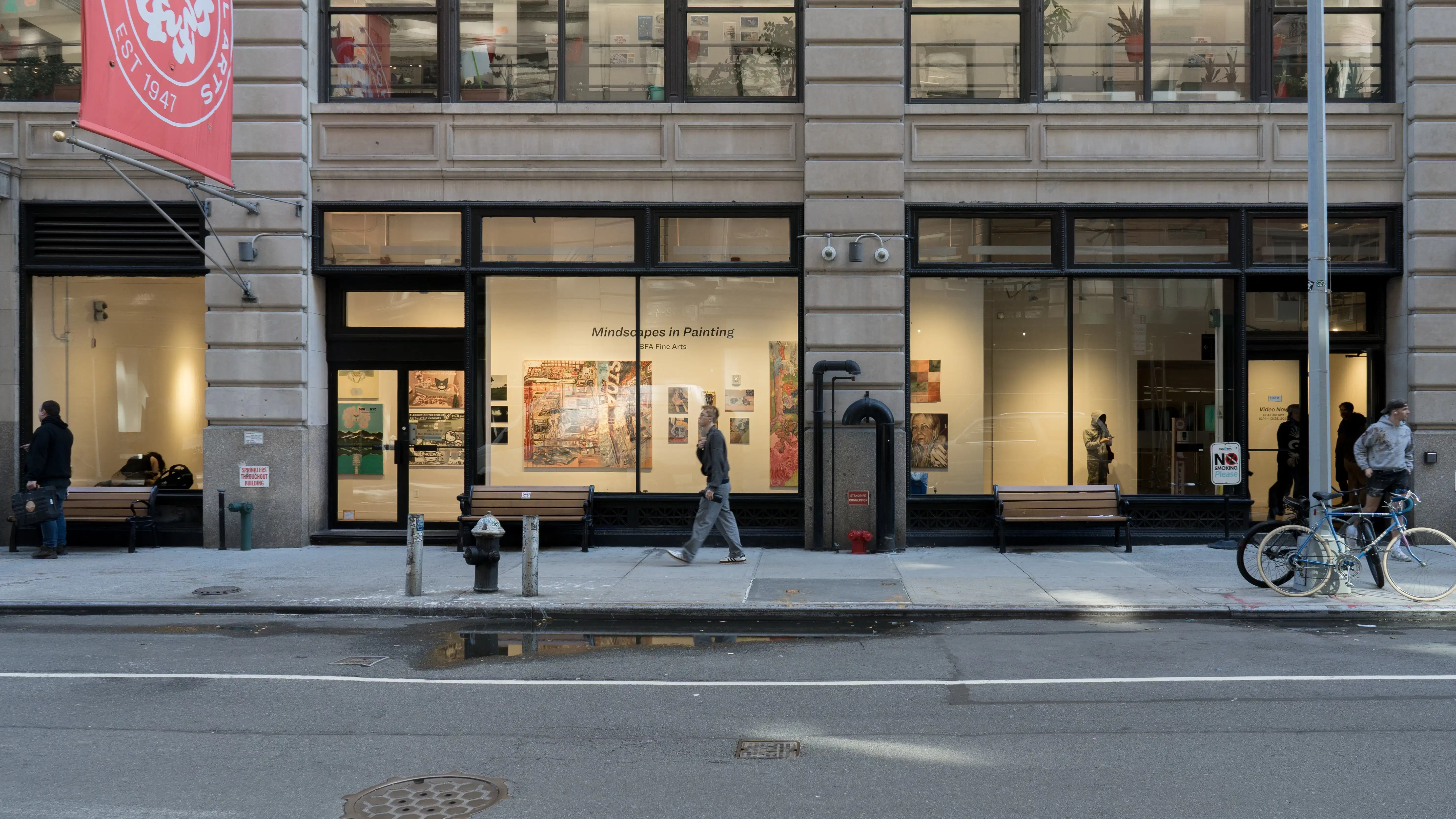 External view of an art gallery inside large picture windows, photographed from across a street, as pedestrians walk down the sidewalk running parallel.