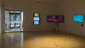 View from inside of gallery space looking toward external glass doors, with three vertically hung monitors to the right of the doors, two hung end-to-end at the corner of the room, and one to its right, each playing video art.