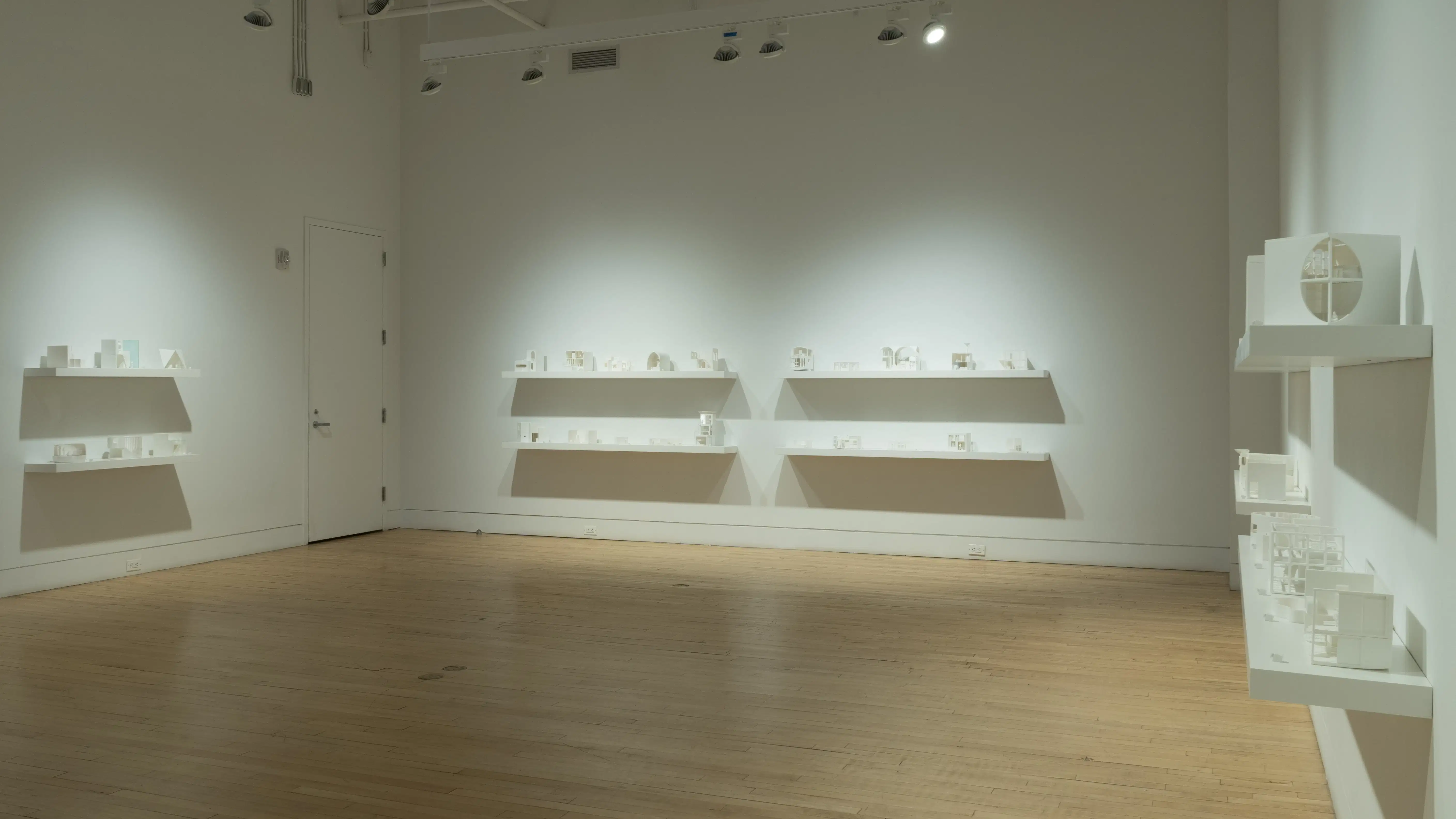 Many wall-mounted shelves containing small white 3D-printed living spaces span three walls of an airy gallery space, stacked in two rows per set. 