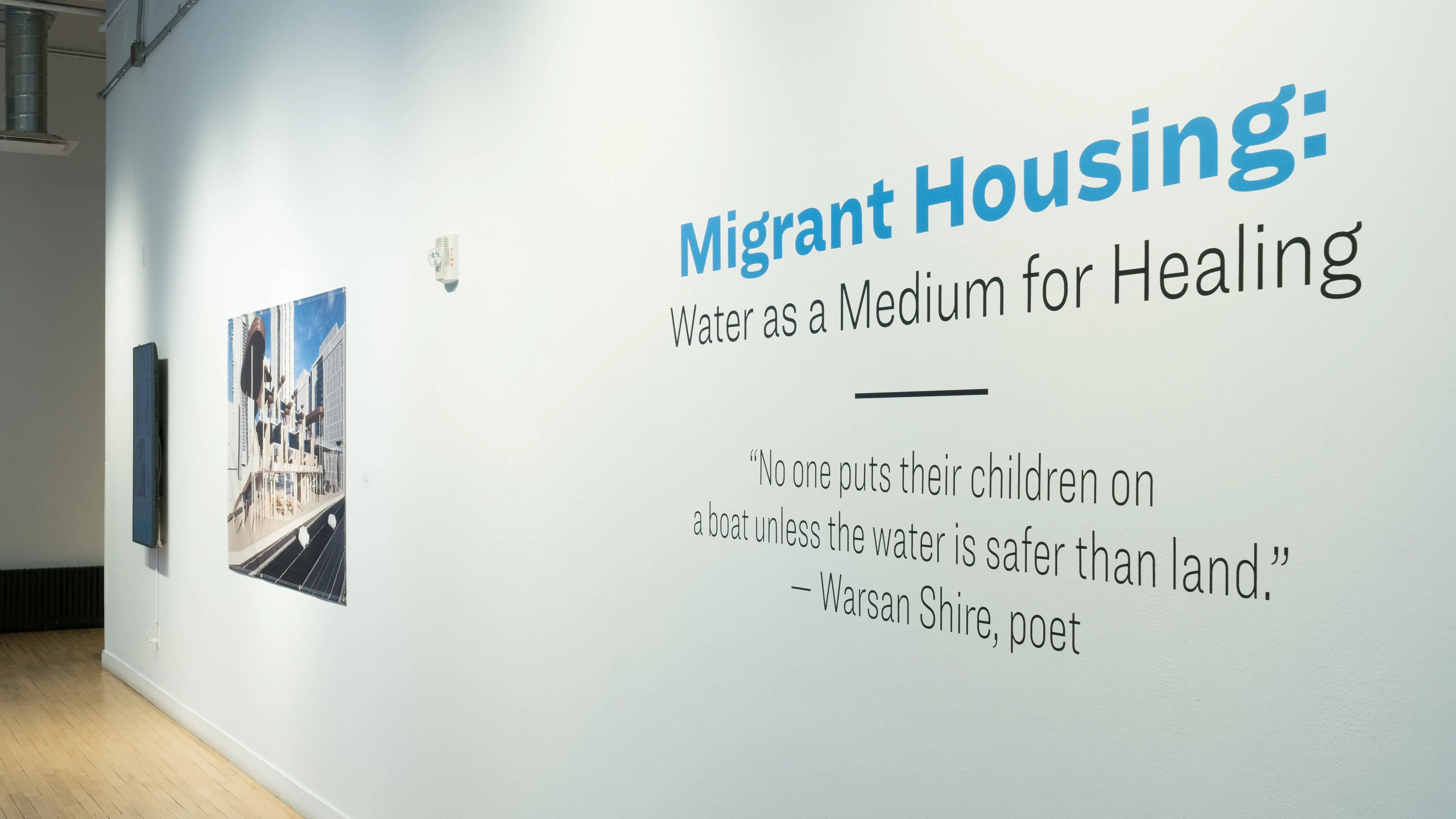 Angled photograph of a long wall displaying the exhibition's title and a quote by Warson Shire, poet, stating "No one puts their children on a boat unless the water is safer than land." To the left of this vinyl text is a large full-color print of a rendered environment and a vertically installed screen.