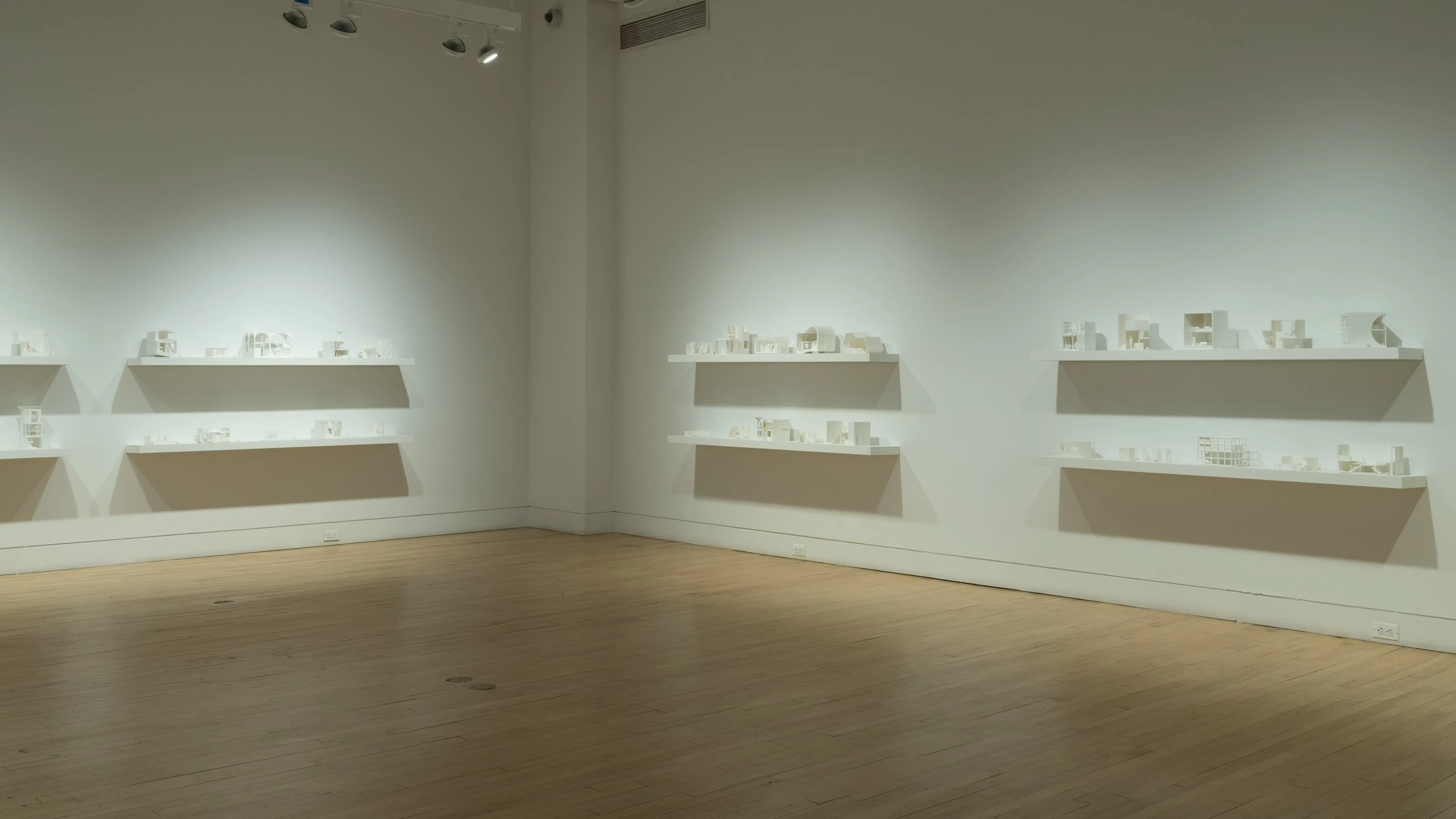 Eight long wall-mounted shelves containing small white 3D-printed living spaces line two walls of a gallery space, stacked in two rows per set. 