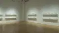 Eight long wall-mounted shelves containing small white 3D-printed living spaces line two walls of a gallery space, stacked in two rows per set. 