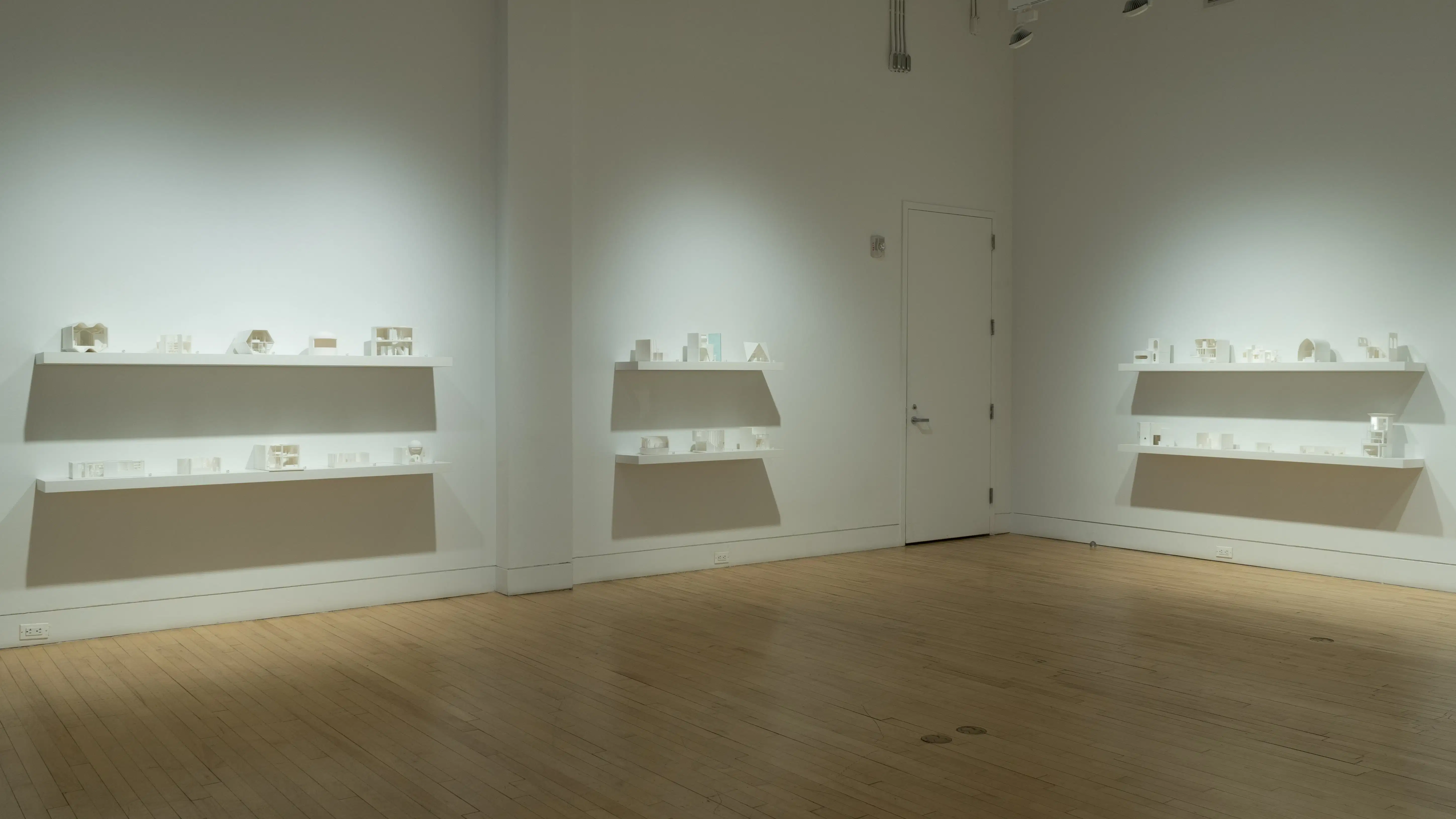 Many wall-mounted shelves containing small white 3D-printed living spaces span three walls of an airy gallery space, stacked in two rows per set. 