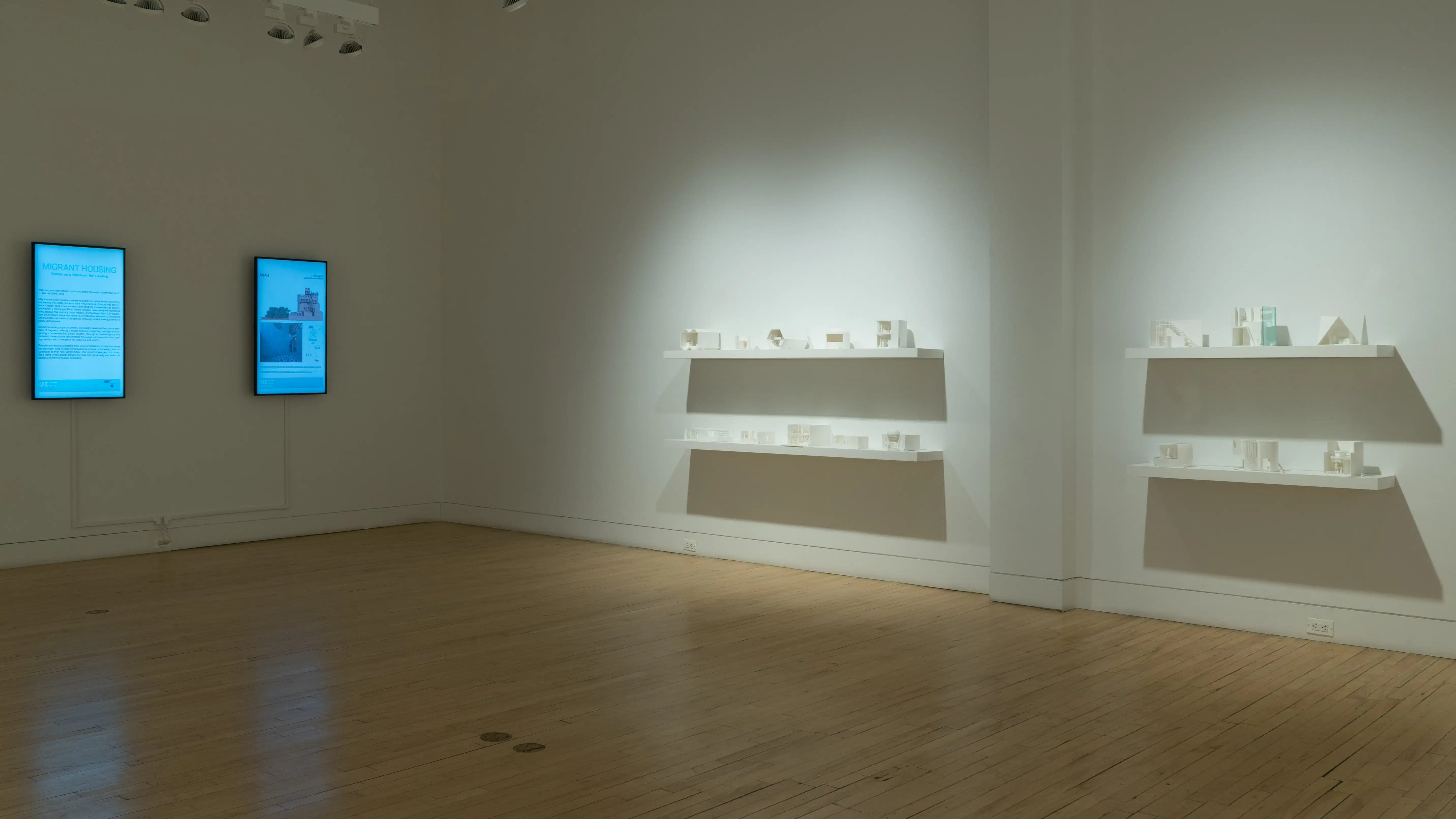 On the far left, two vertical monitors display graphics about the two colleges' programs and additional visualizations of student projects. On the right are four long shelves, stacked in groups of two, displaying small 3D-printed iterations of student designs. 