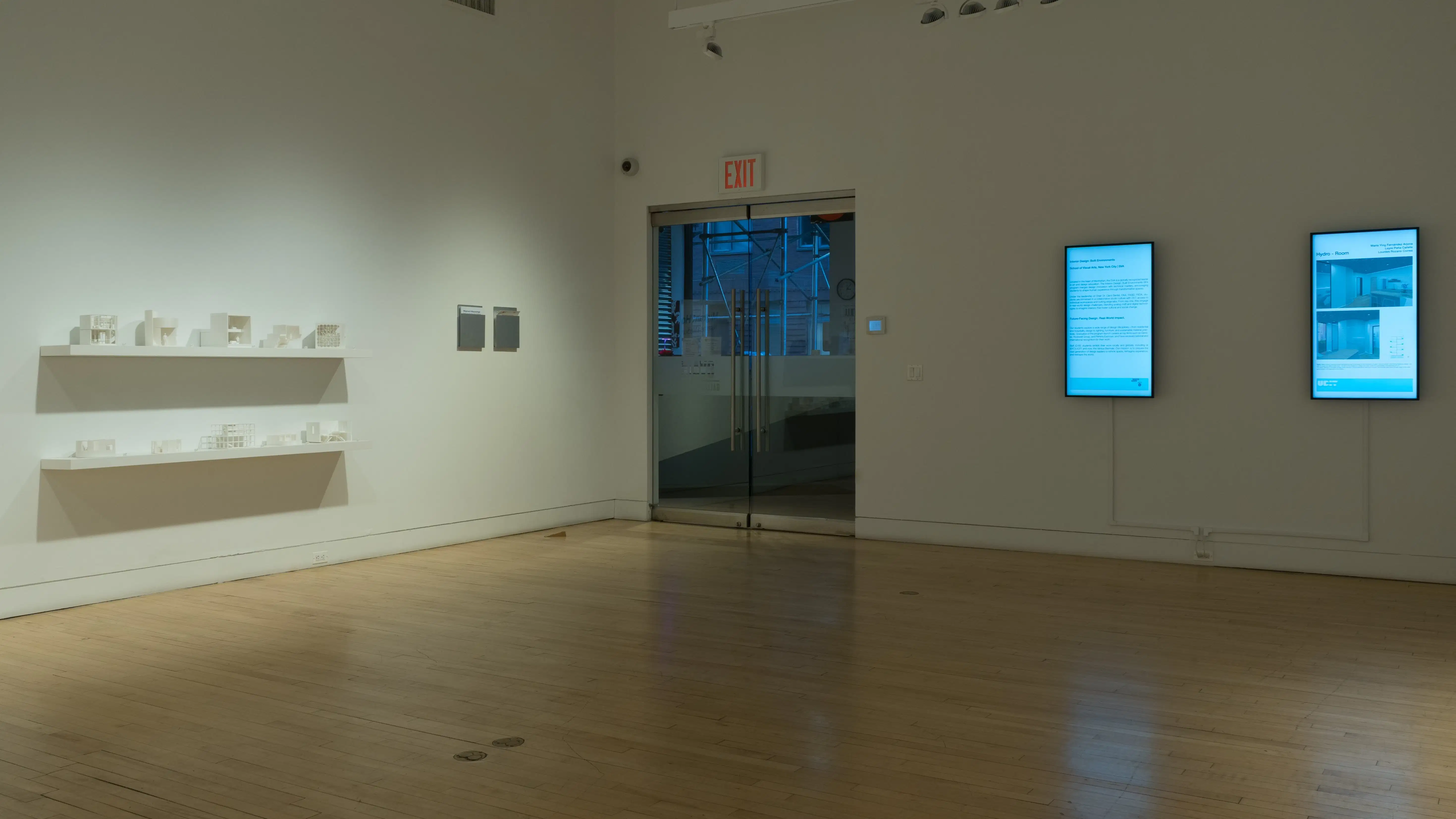 On the far left, two shelves stacked vertically display many small 3D-printed iterations of student work. In the center of the photo is a double glass-door exit, and to its right are two vertically oriented monitors displaying information about the two colleges' programs and additional student design work.