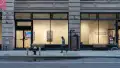 From the outside, the SVA Flatiron Gallery is visible through the building's windows. Two monitors, one horizontal and one vertical, are on display in the hallway behind the windows. On the right side of the wall, vinyl text reads “Technological Perseverance.”