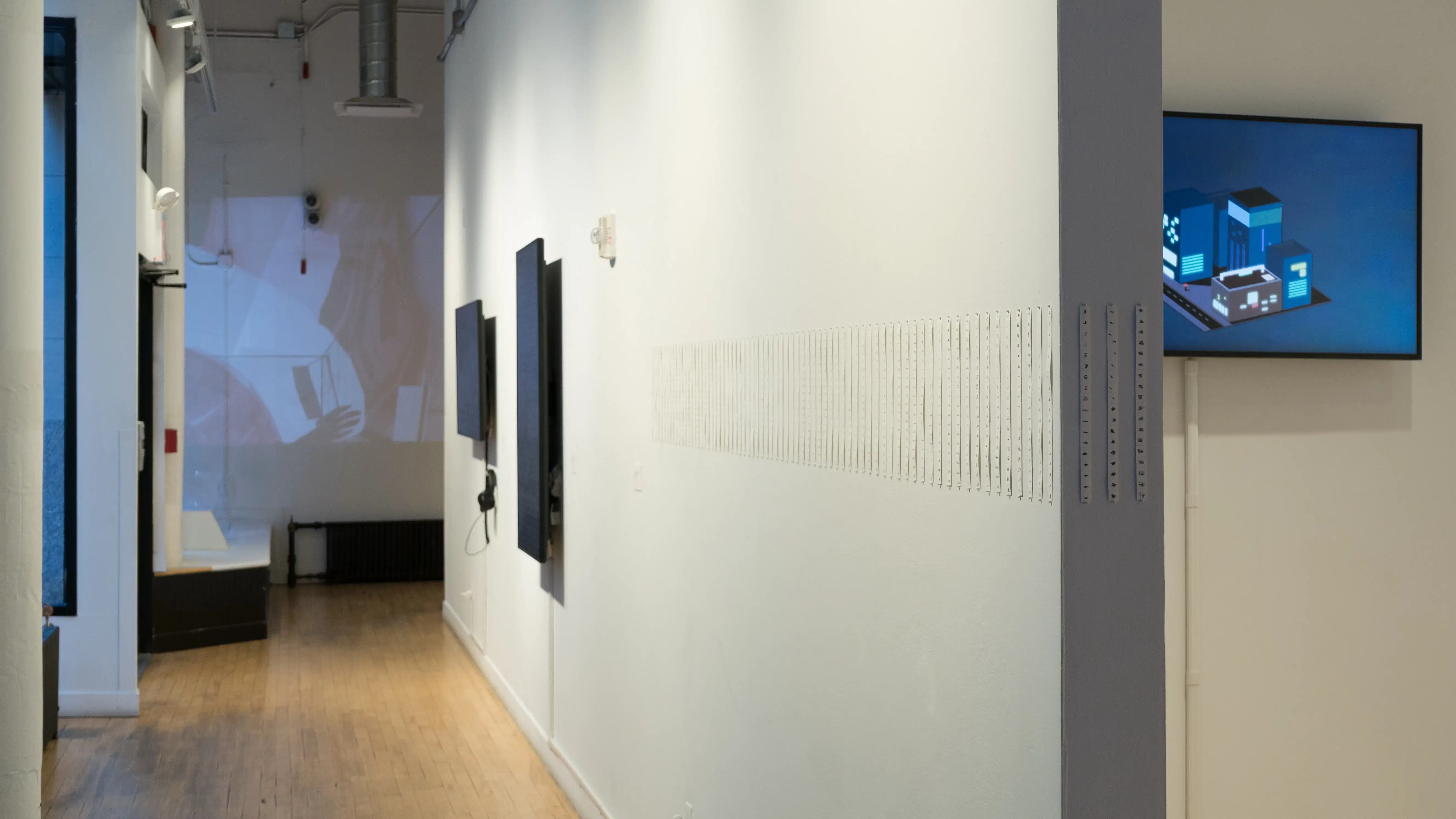 Looking down the hallway in front of the building windows, thin strips of paper are displayed in a sequence. Beyond them, there are two monitors on the wall and a projection at the end of the hall.