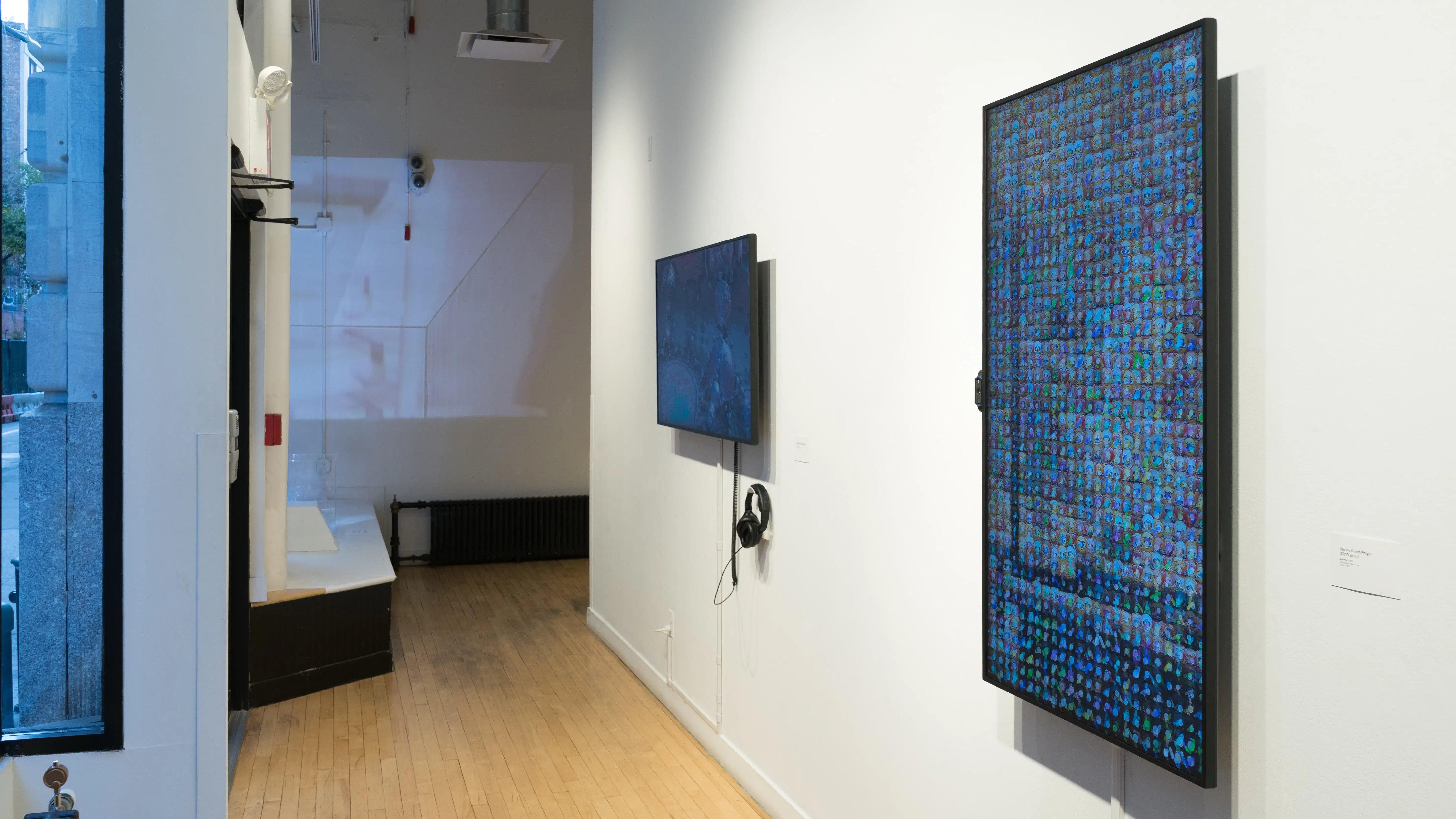 Towards the end of the hallway, a vertical monitor on the wall shows many small square images arranged all together. To its left, a horizontal monitor plays something else, and a projection on the far end shows a third thing.