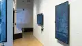 Towards the end of the hallway, a vertical monitor on the wall shows many small square images arranged all together. To its left, a horizontal monitor plays something else, and a projection on the far end shows a third thing.