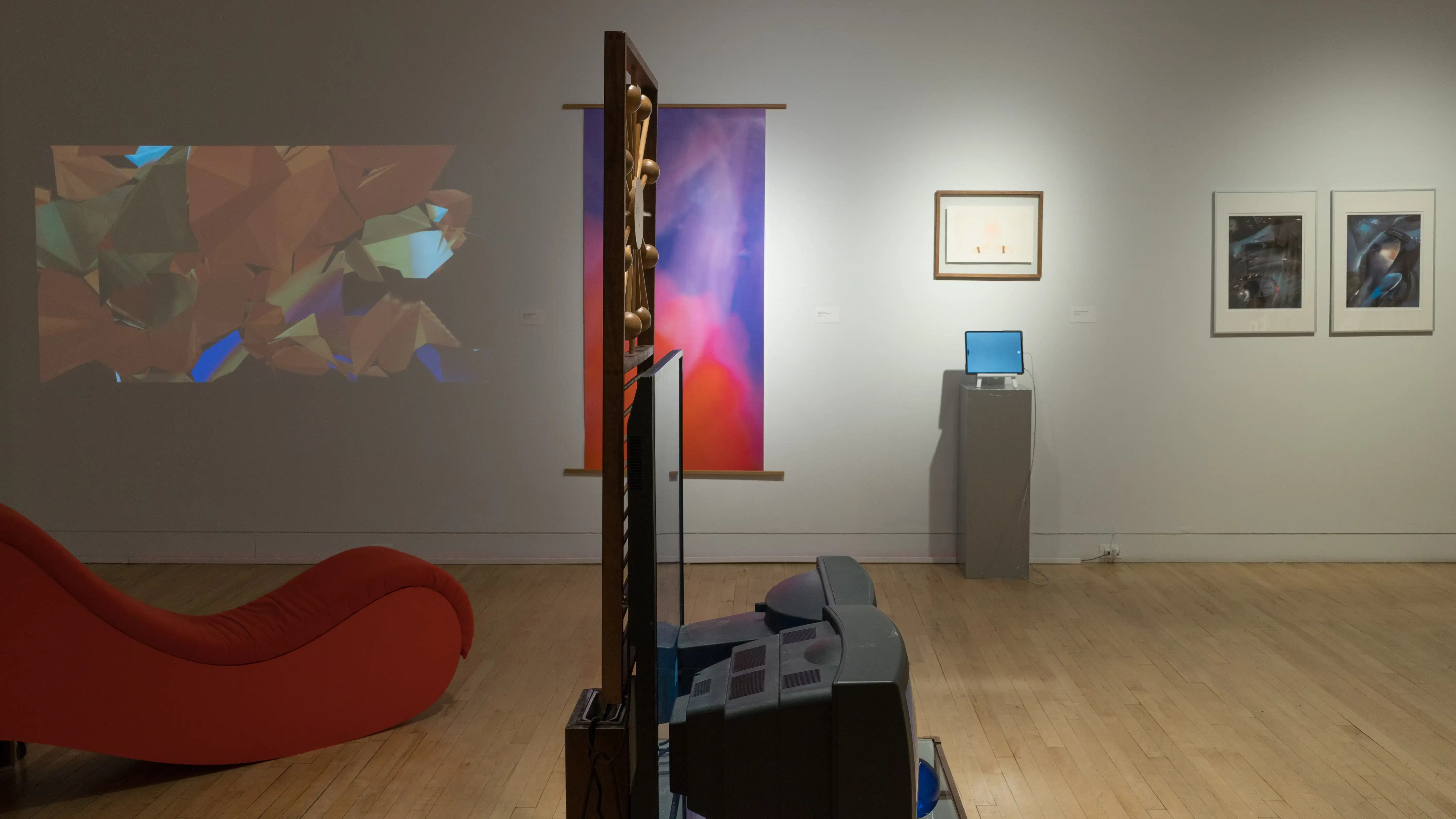 Looking past the installation in the center of the room, a red retro-looking chair is placed before the projection. To the right of the projection is a long vertical print of an abstract pink, red, and purple design. Beyond that, framed prints hang on the wall above and beside an iPad resting on a pedestal.