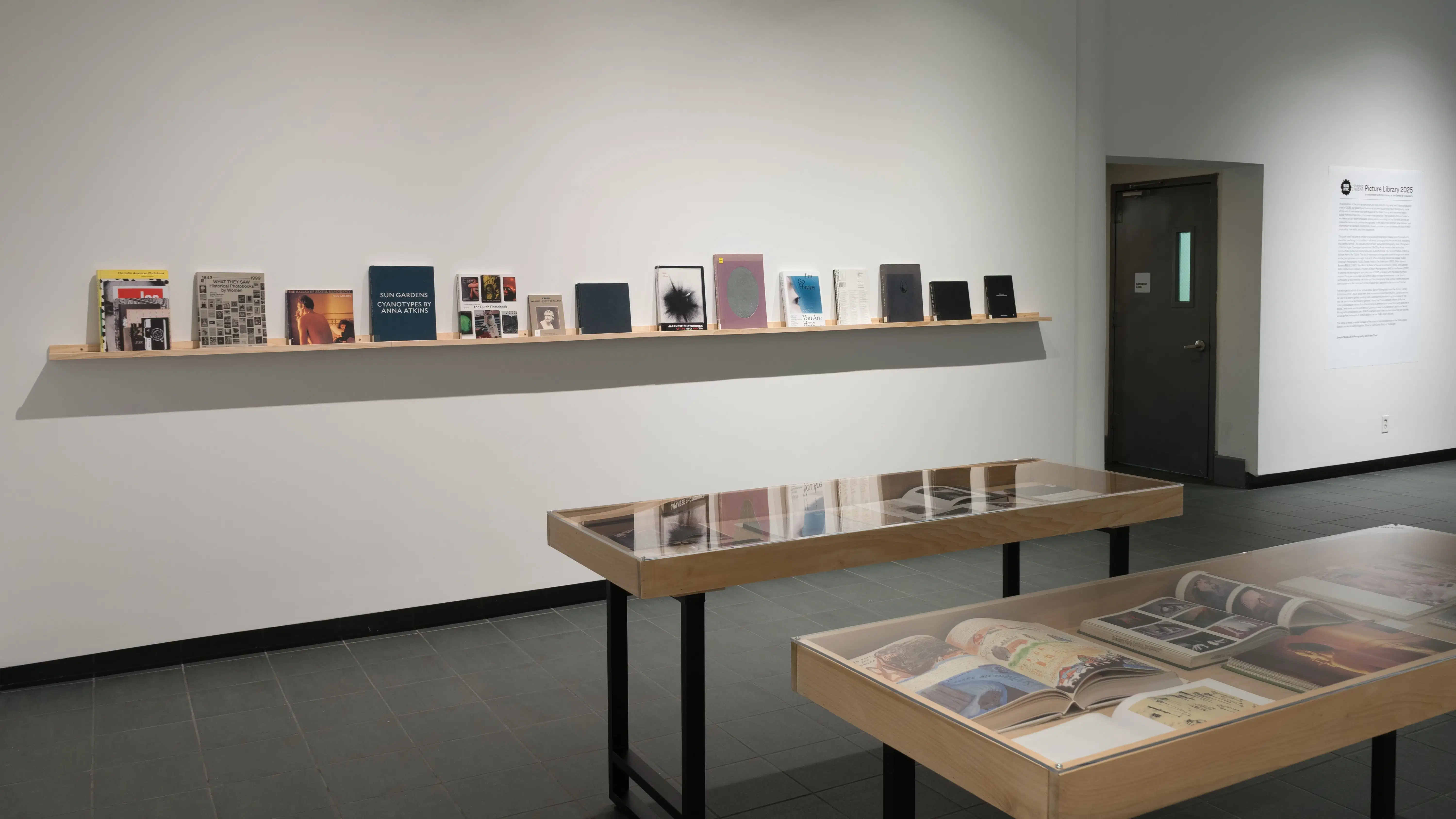 Two small vitrines stand in the center of the SVA Gramercy Gallery lobby. Behind them, one long wooden shelf runs down a wall. Many front-facing books of various sizes sit on the shelf.