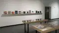 Two small vitrines stand in the center of the SVA Gramercy Gallery lobby. Behind them, one long wooden shelf runs down a wall. Many front-facing books of various sizes sit on the shelf.