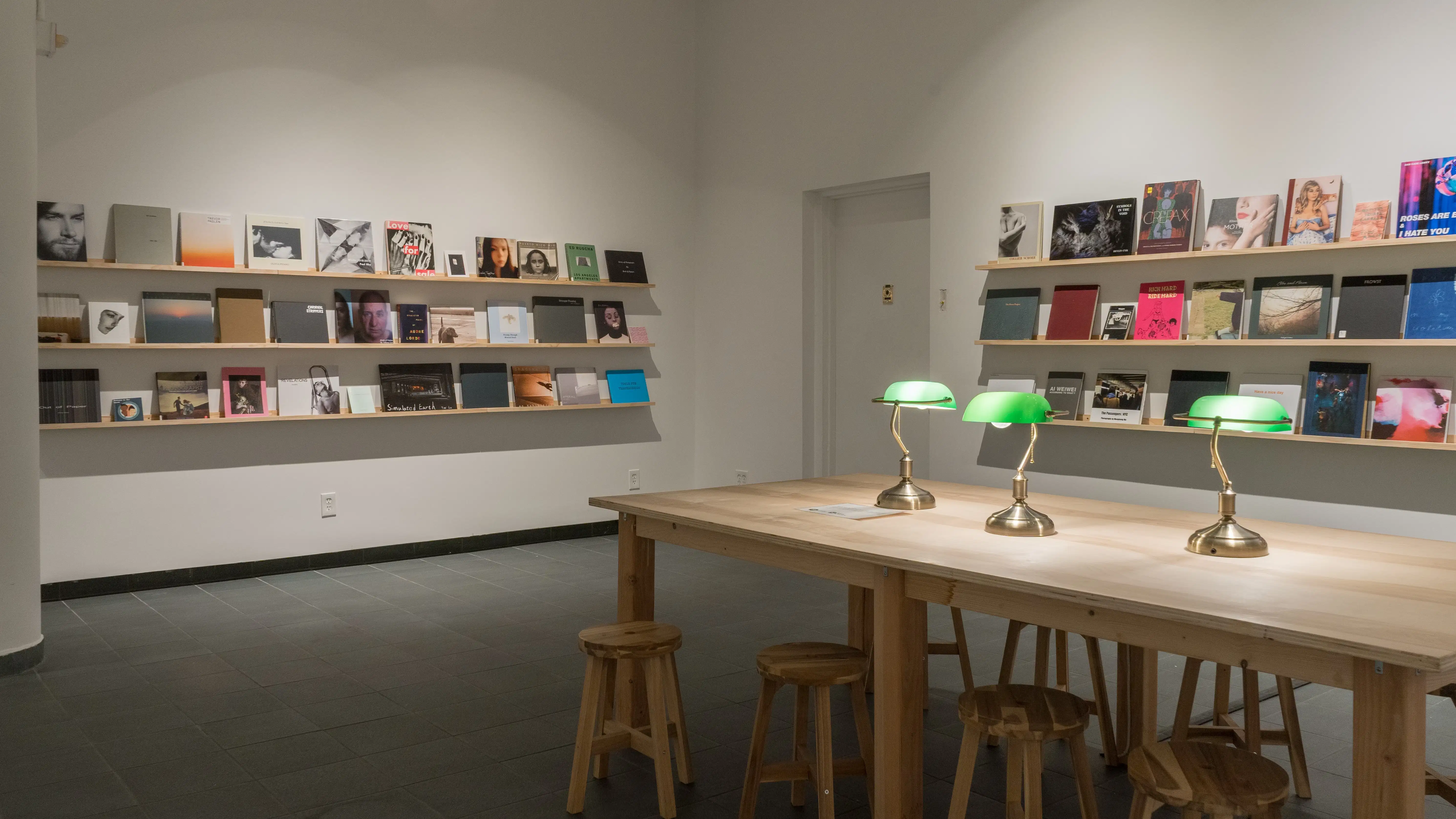 Each of the two visible walls is lined with three long, wooden shelves. Front-facing books of different sizes, colors, and designs fill all of the shelves. A large wooden table, accompanied by wooden stools and green table lamps, is set up in the center of the room.