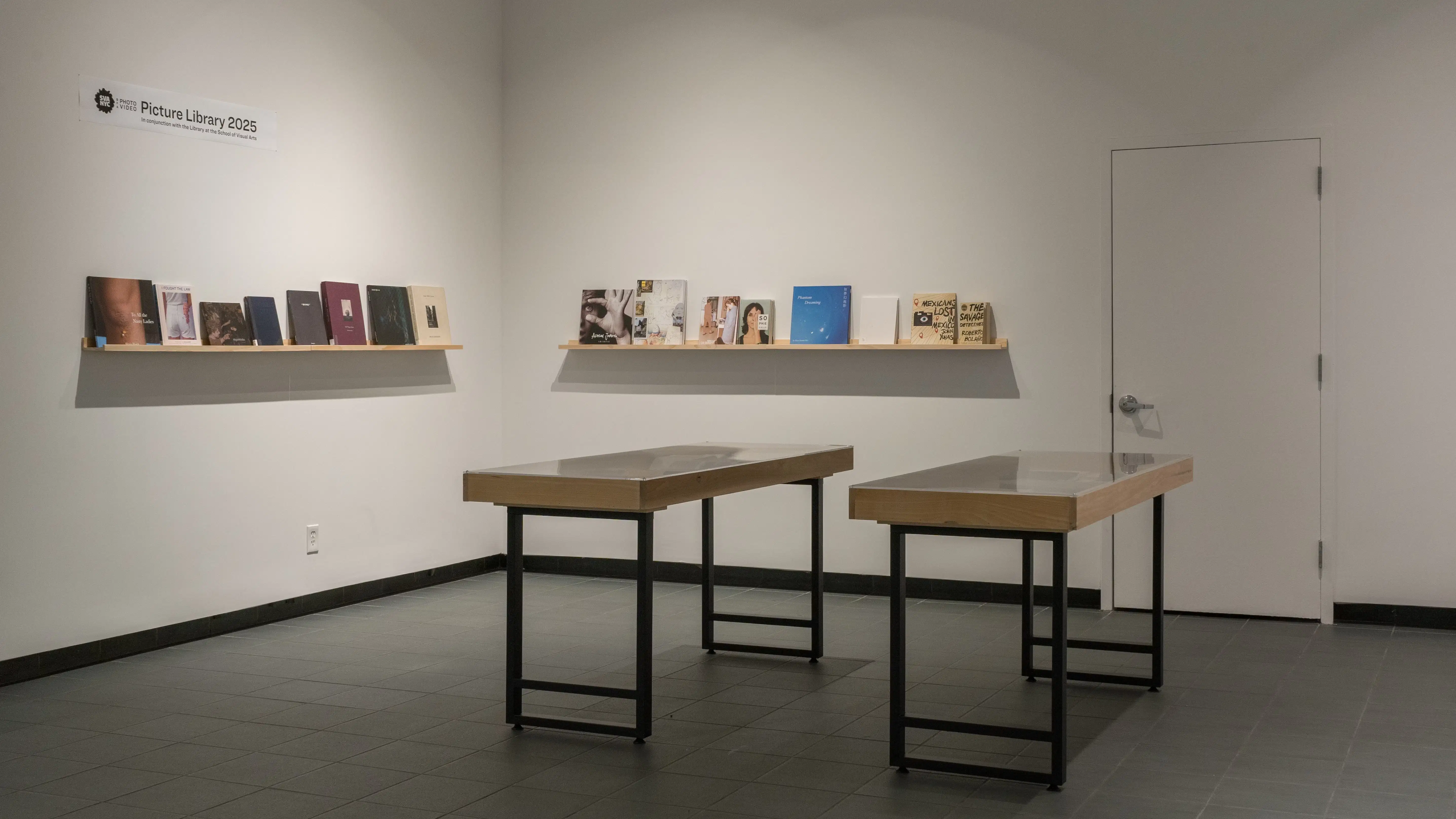 Two small vitrines stand in the center of the SVA Gramercy Gallery lobby. Behind them, two walls each have a single wooden shelf holding front-facing books of various sizes. A small sign above the shelves reads “Picture Library 2025.”