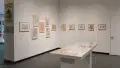 In the lobby of the SVA Gramercy Gallery, framed illustrations by Lillian Ansel are displayed on the walls. In the center of the floor, a vitrine holds loose papers.