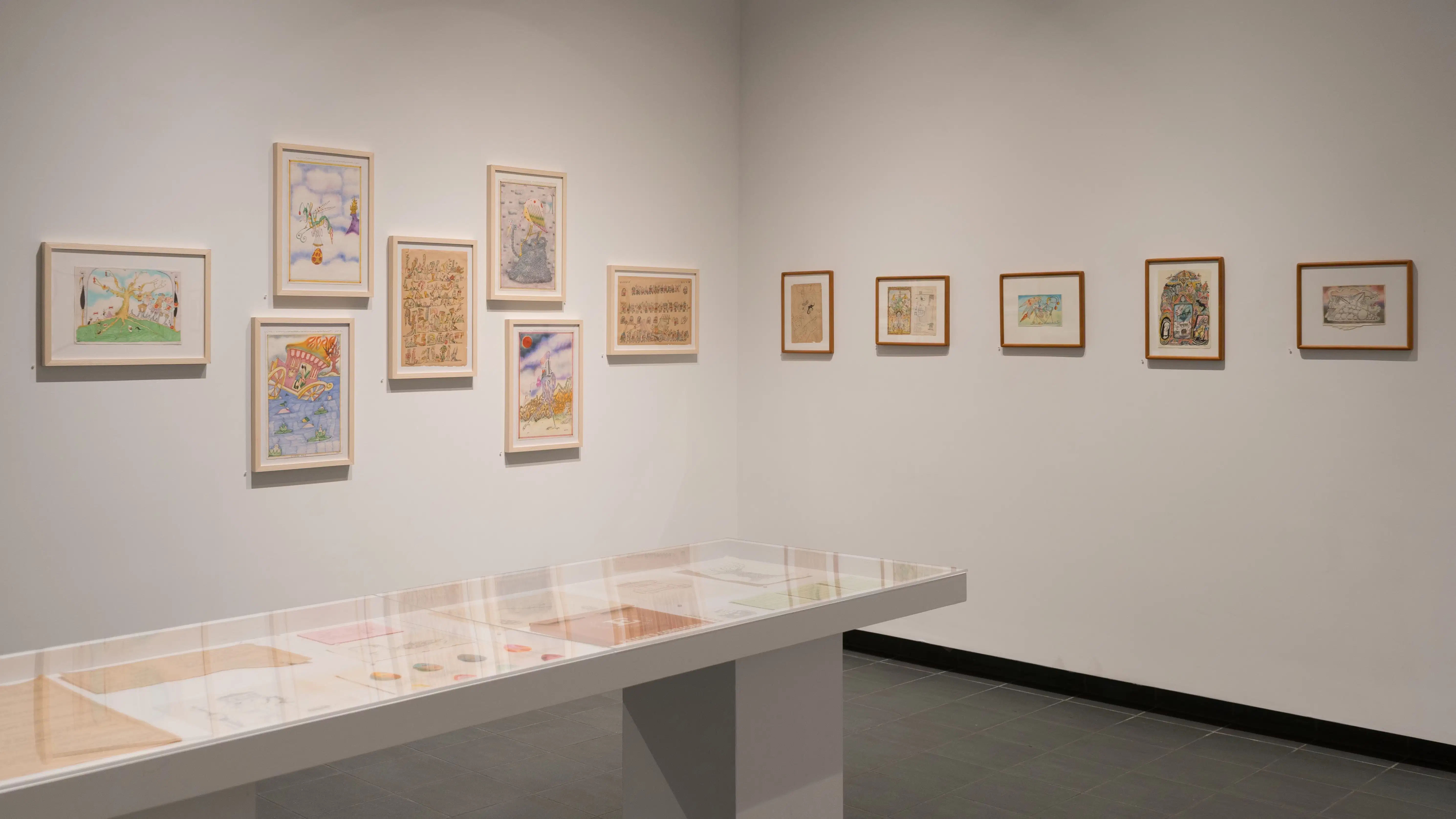 Framed illustrations by Lillian Ansel are displayed on the walls. A vitrine, in front of the walls, holds loose papers. The framed illustrations vary in size and are colorful and whimsical.