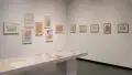 Framed illustrations by Lillian Ansel are displayed on the walls. A vitrine, in front of the walls, holds loose papers. The framed illustrations vary in size and are colorful and whimsical.