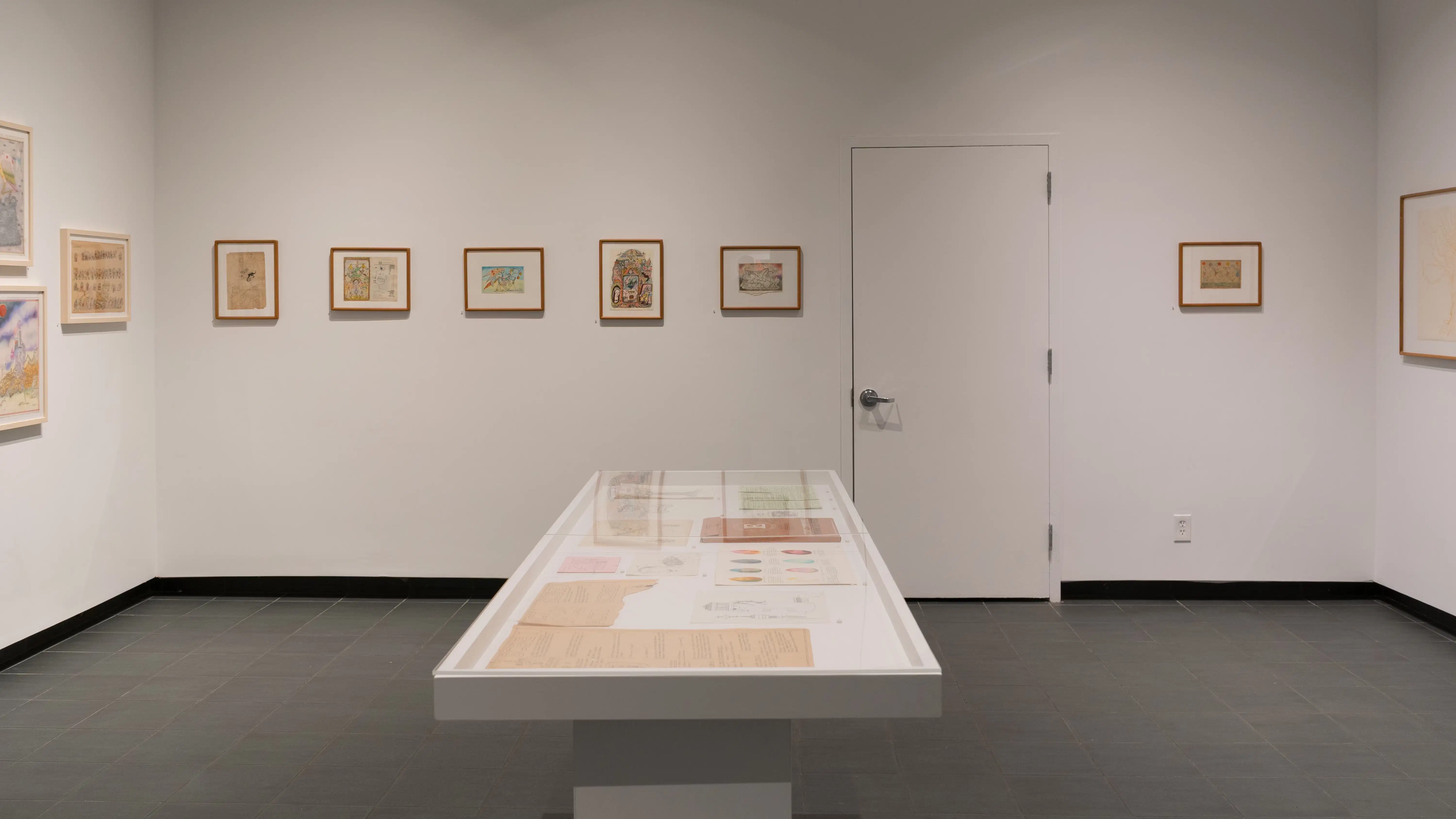 On one wall, small illustrations are framed in wooden frames with white mats. Some are oriented vertically, while others are oriented horizontally. A vitrine, in front of the walls, holds loose papers.