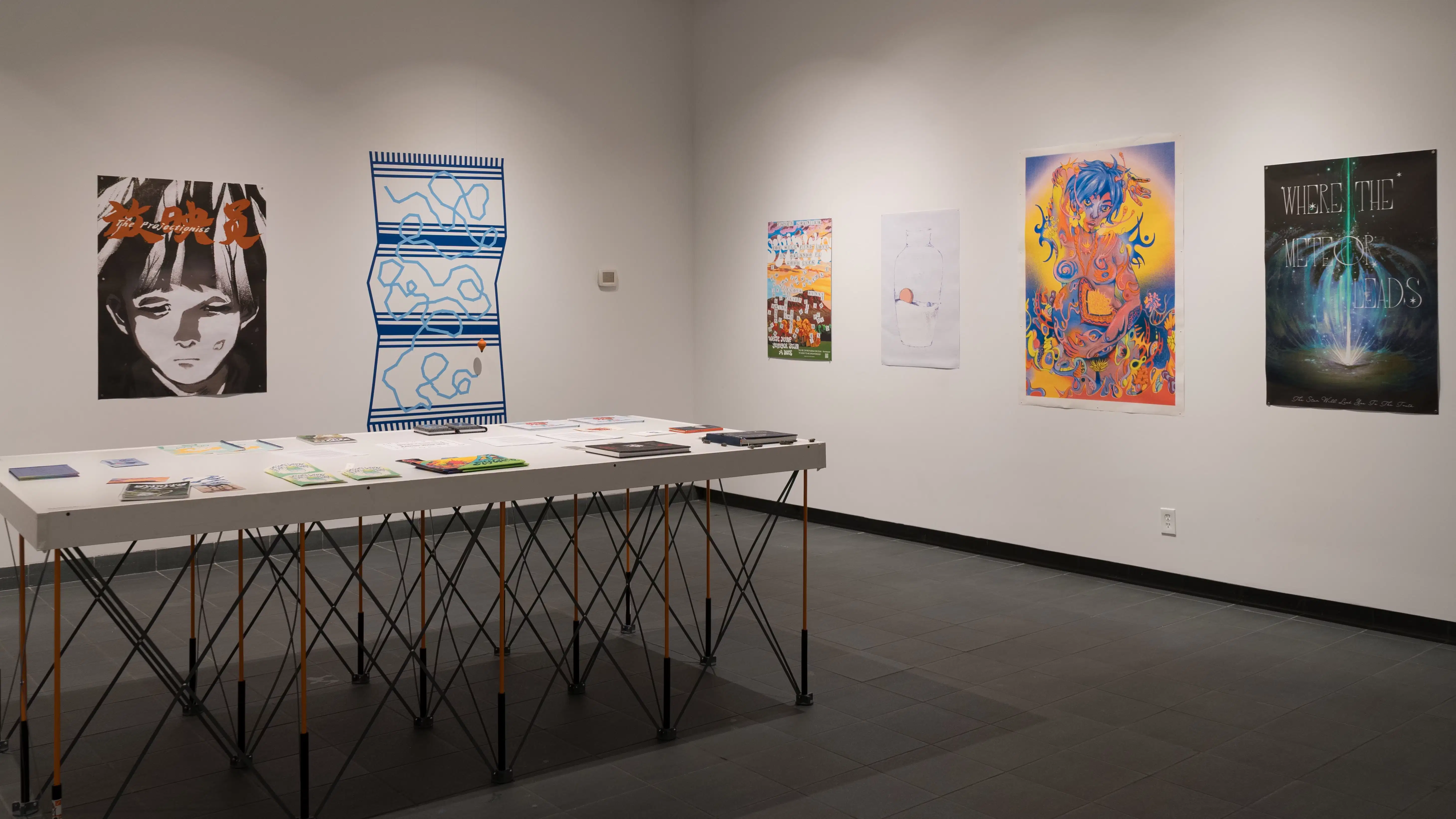 Many books lay flat on a white table near the corner of the room. Beyond the table, prints and vinyl artworks are displayed on the wall.