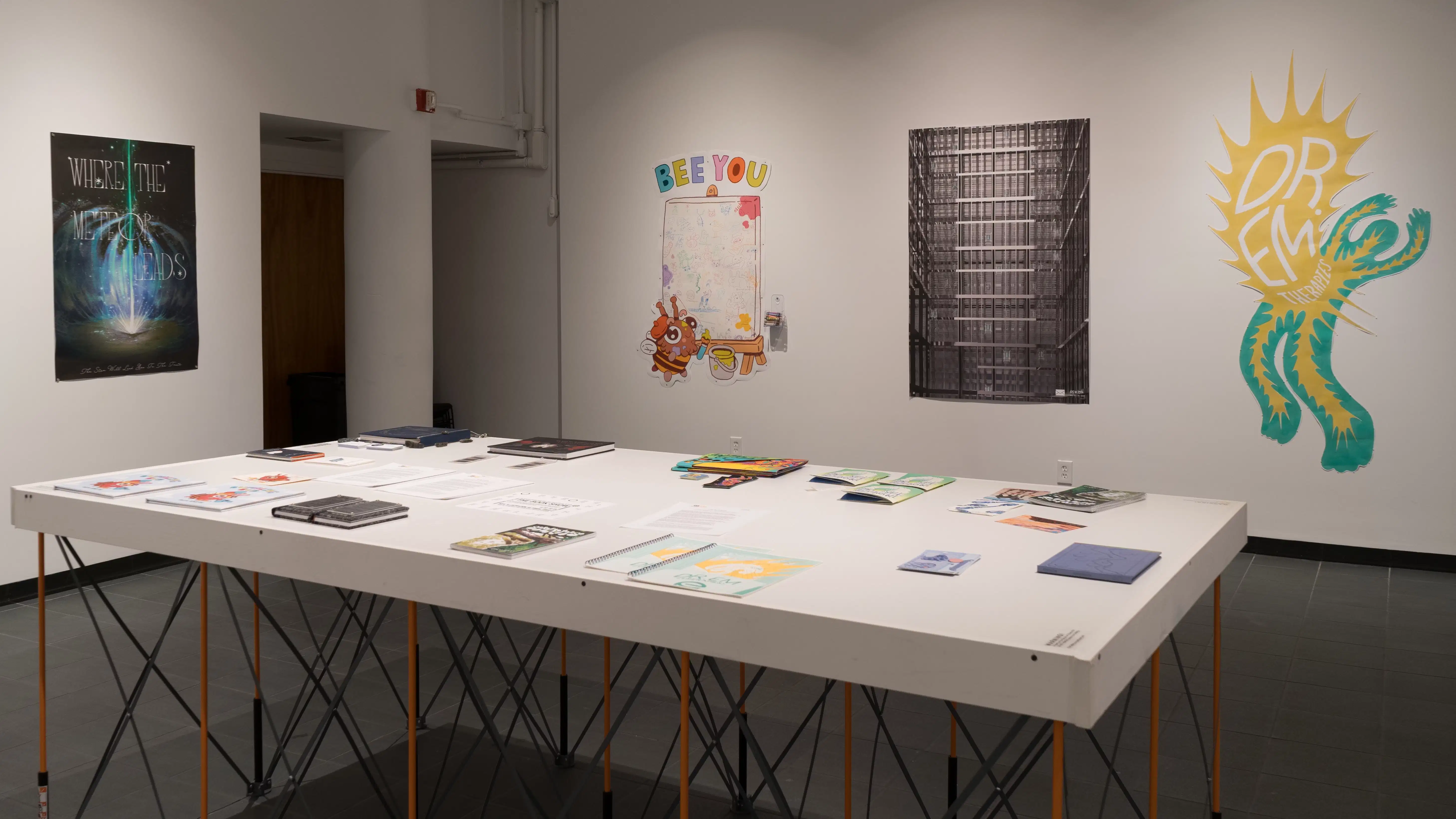 Many books lay flat on a white table near the entrance to the room. Beyond the table, prints and cut-paper artworks are displayed on the wall.